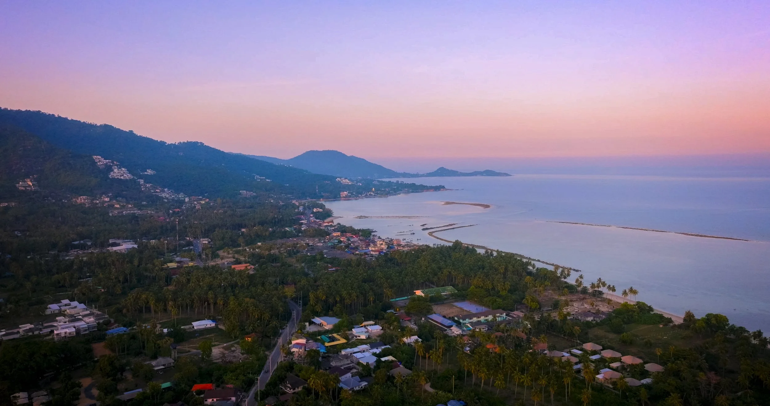 A coastal landscape at sunset with hills, a small town, and a calm sea with a curved jetty.