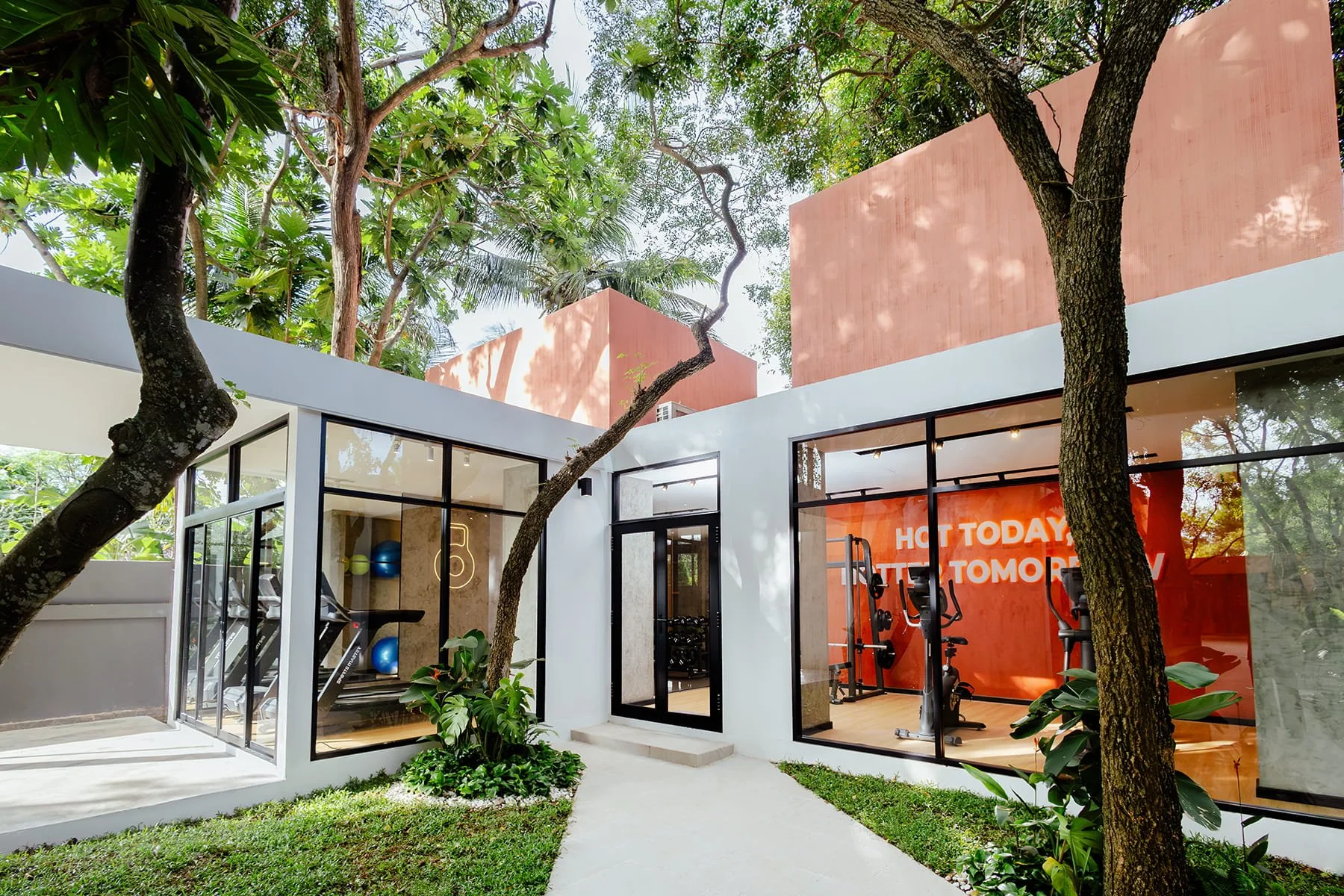 Modern gym building with large glass windows surrounded by trees and greenery, interior with exercise equipment, and a motivational quote on the wall.