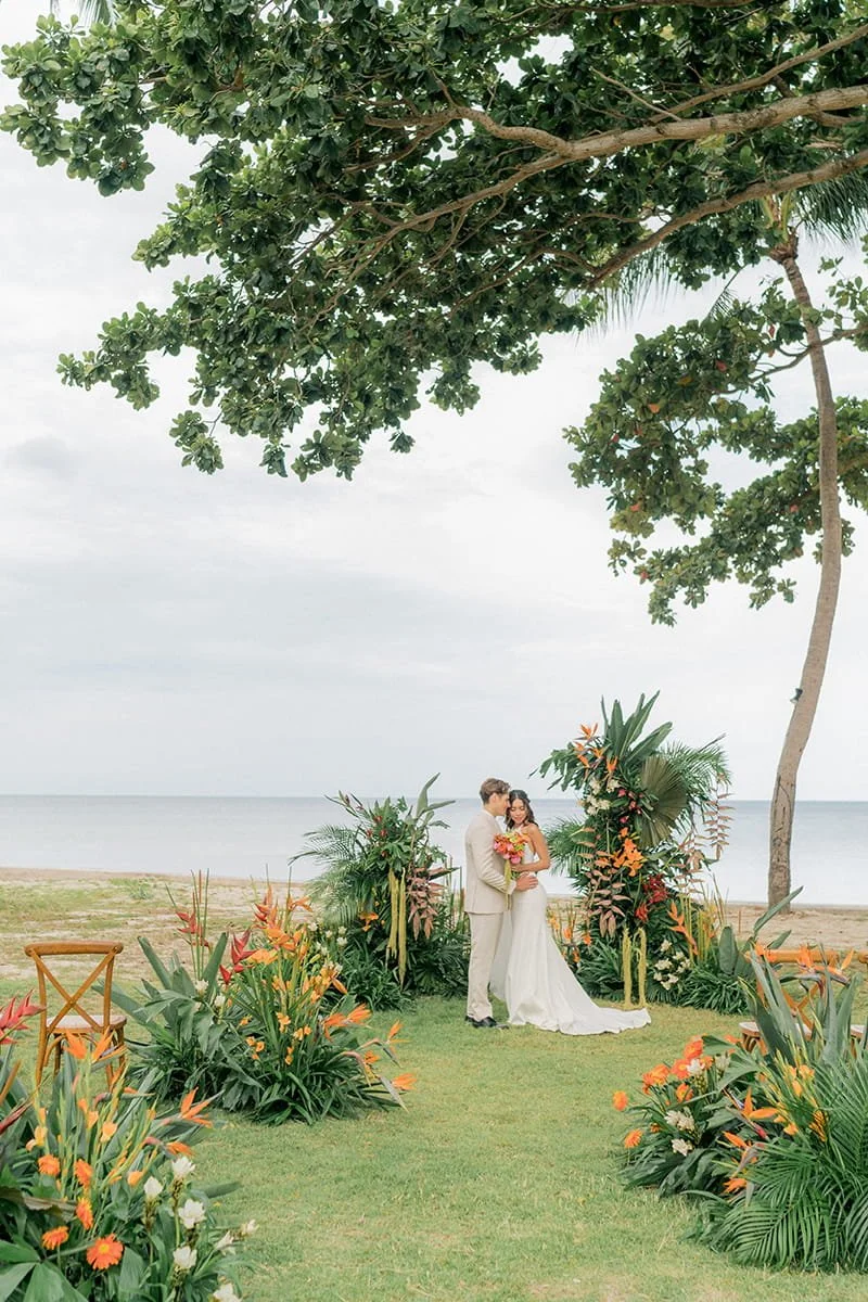 A couple in wedding attire standing under a floral arch on a grassy area by the beach, surrounded by tropical plants and flowers, with beach and ocean in the background.