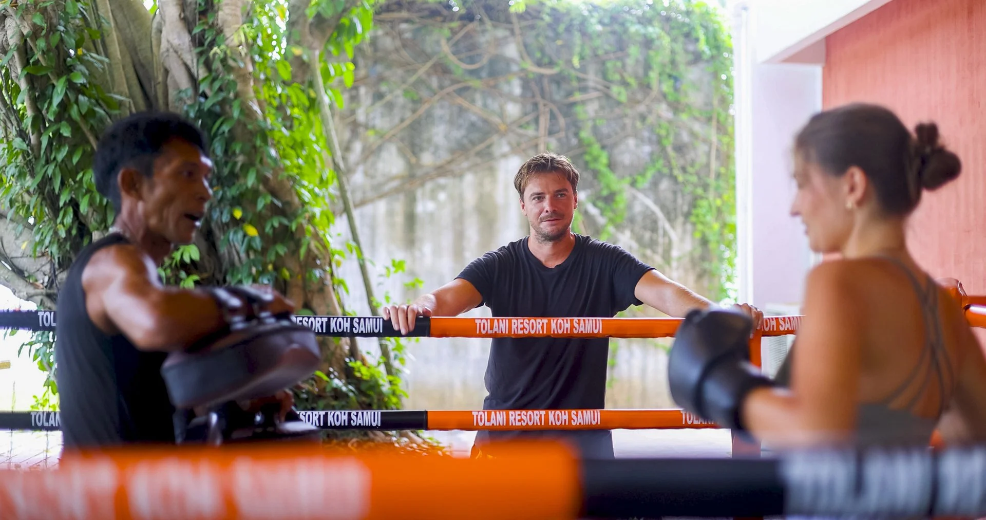 A boxing or Muay Thai training session with a male trainer standing behind orange and black ropes labeled 'TOLANI RESORT KOH SAMUI,' observing two female fighters wearing black gloves and sports bras, with a lush outdoor green background.