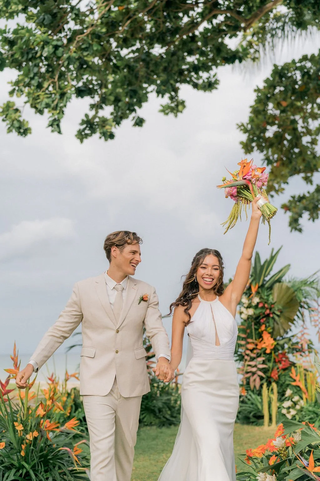 A joyful bride and groom walking hand in hand outdoors, the bride holding a colorful bouquet raised in the air, surrounded by lush tropical plants and greenery.
