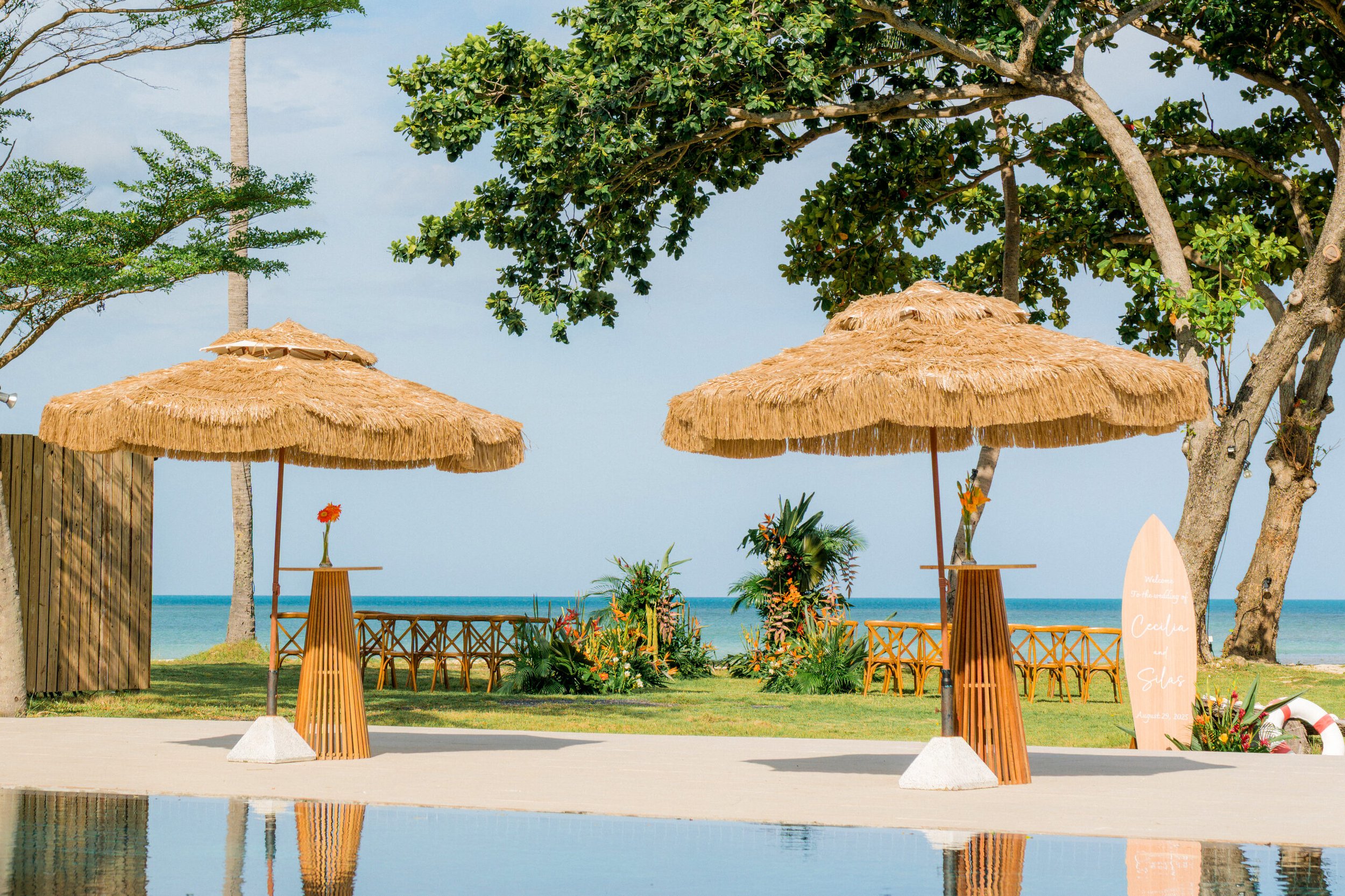 Two straw beach umbrellas with small tables holding flowers, set up near a swimming pool on a beach with palm trees and an ocean view in the background.