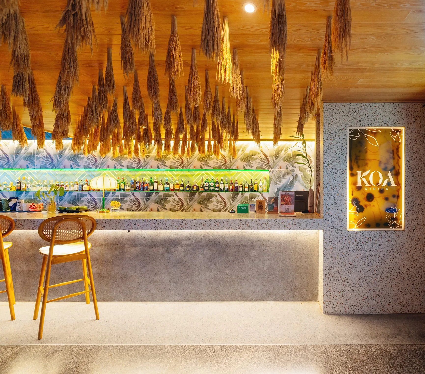 Modern bar with a terrazzo counter, tropical wallpaper, hanging dried plants, and a backlit sign with 'KOA' and 'Dining' on a decorative panel.
