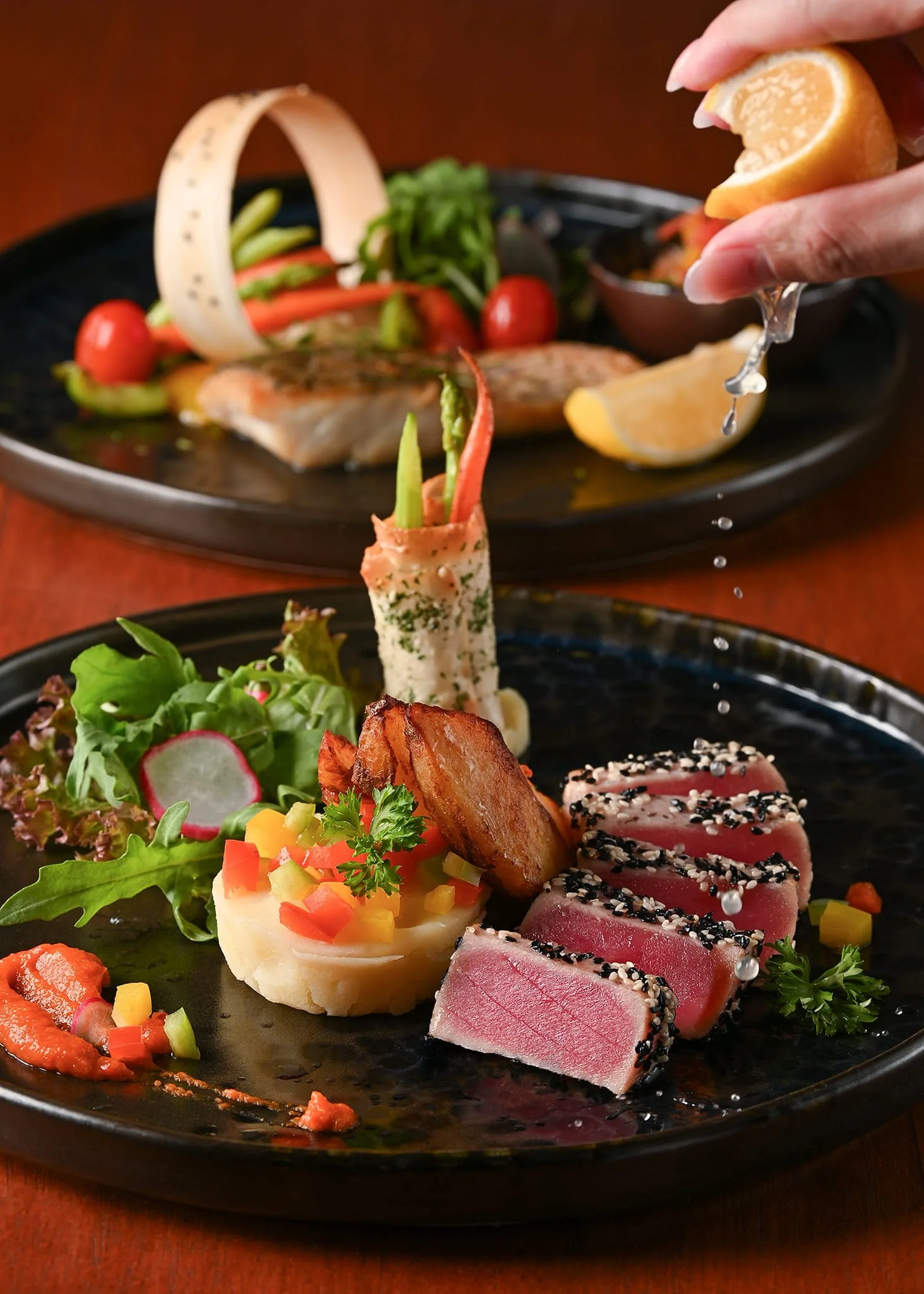 Close-up of a plated meal featuring seared tuna with black sesame seeds, a potato salad with diced vegetables, a grilled potato wedge, and a mixed green salad with radish slices on a black plate. In the background, a person garnishing the plate with 