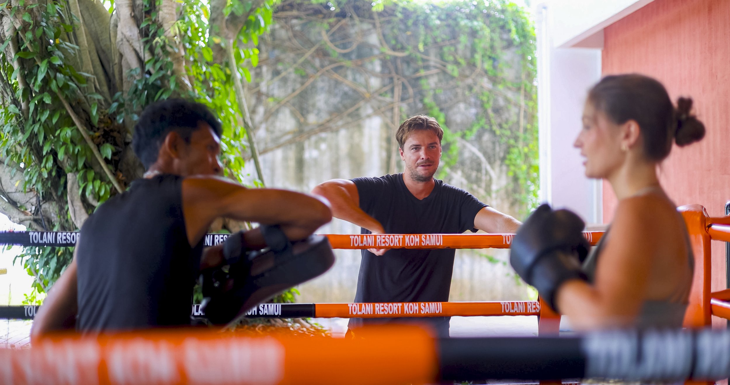 A fitness trainer is coaching two boxers in a boxing ring. The trainer, in a black shirt, is assisting a woman with boxing gloves on, while another man watches nearby. The ring is outdoors with green trees and a rocky background, and orange ropes sur