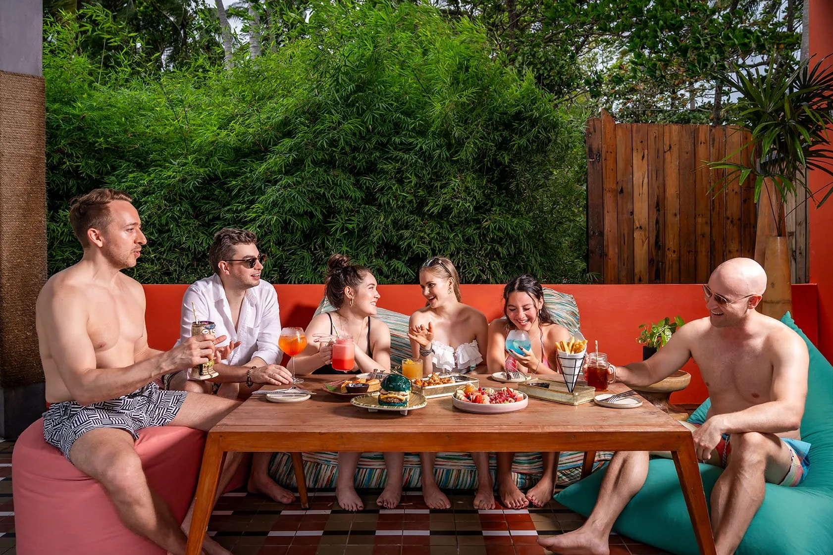 Group of six friends sitting around a table outdoors, smiling, drinking colorful beverages, and enjoying snacks and desserts.