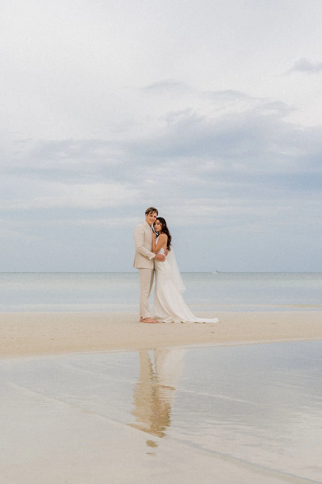 A couple in wedding attire standing on a beach with a cloudy sky and the ocean in the background, their reflection visible in the water.