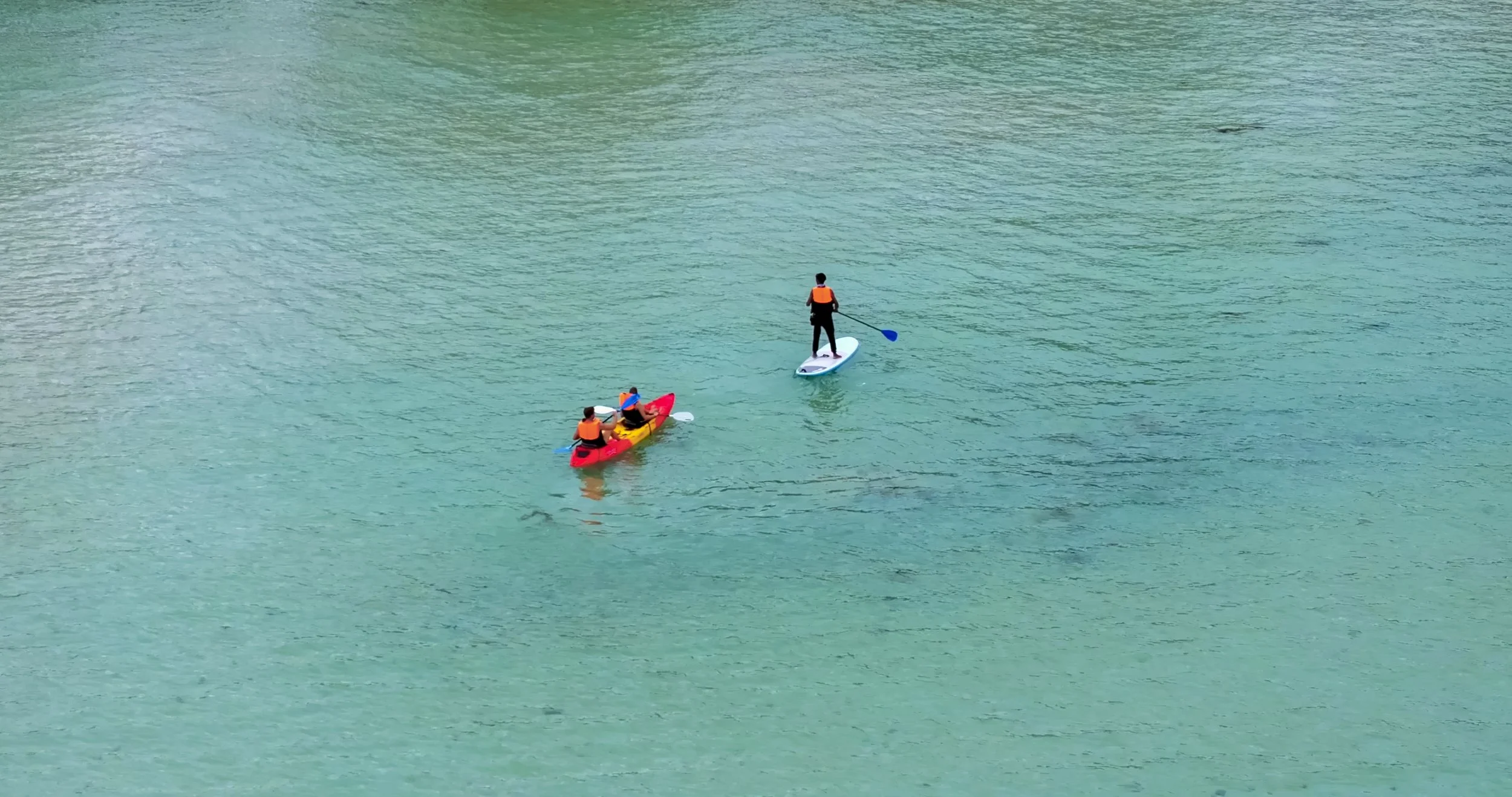 Three people paddleboarding on a calm, turquoise lake, wearing orange life jackets.