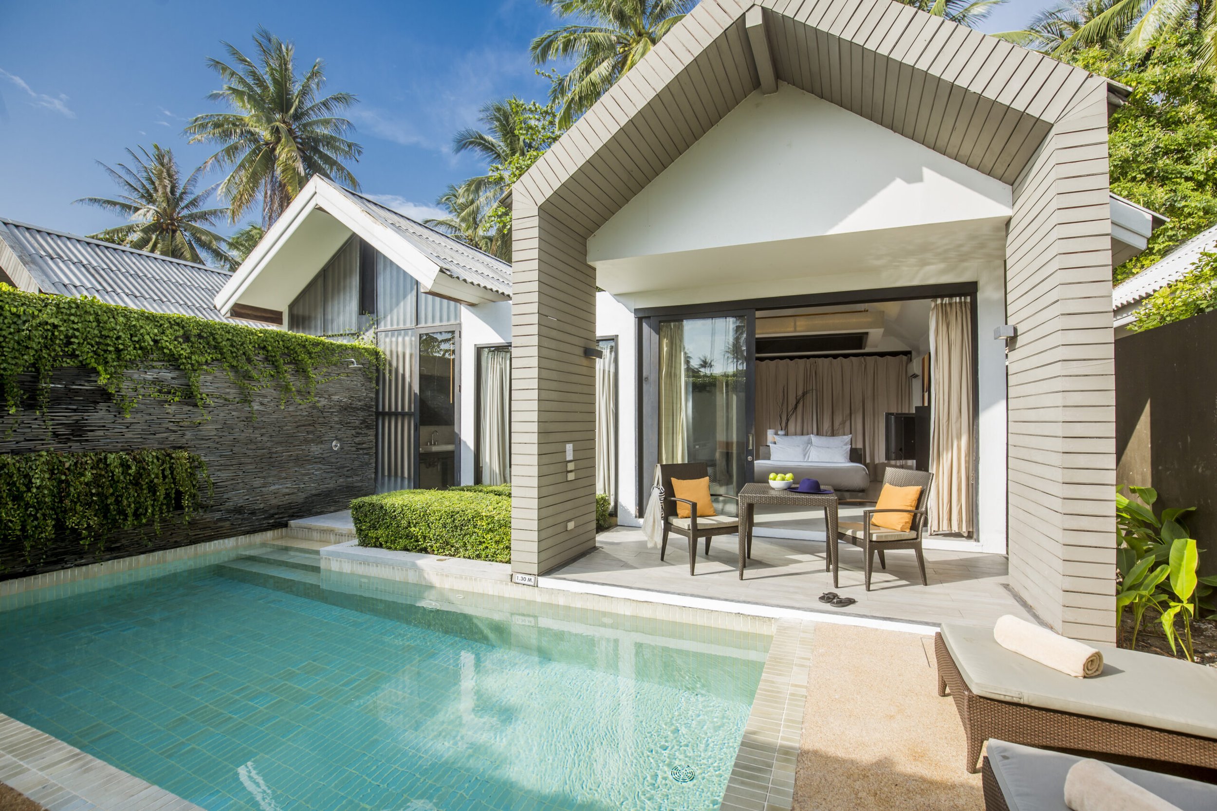 Luxury villa with a swimming pool, outdoor patio with chairs and table, and a bedroom with a view through sliding glass doors, surrounded by tropical palm trees.