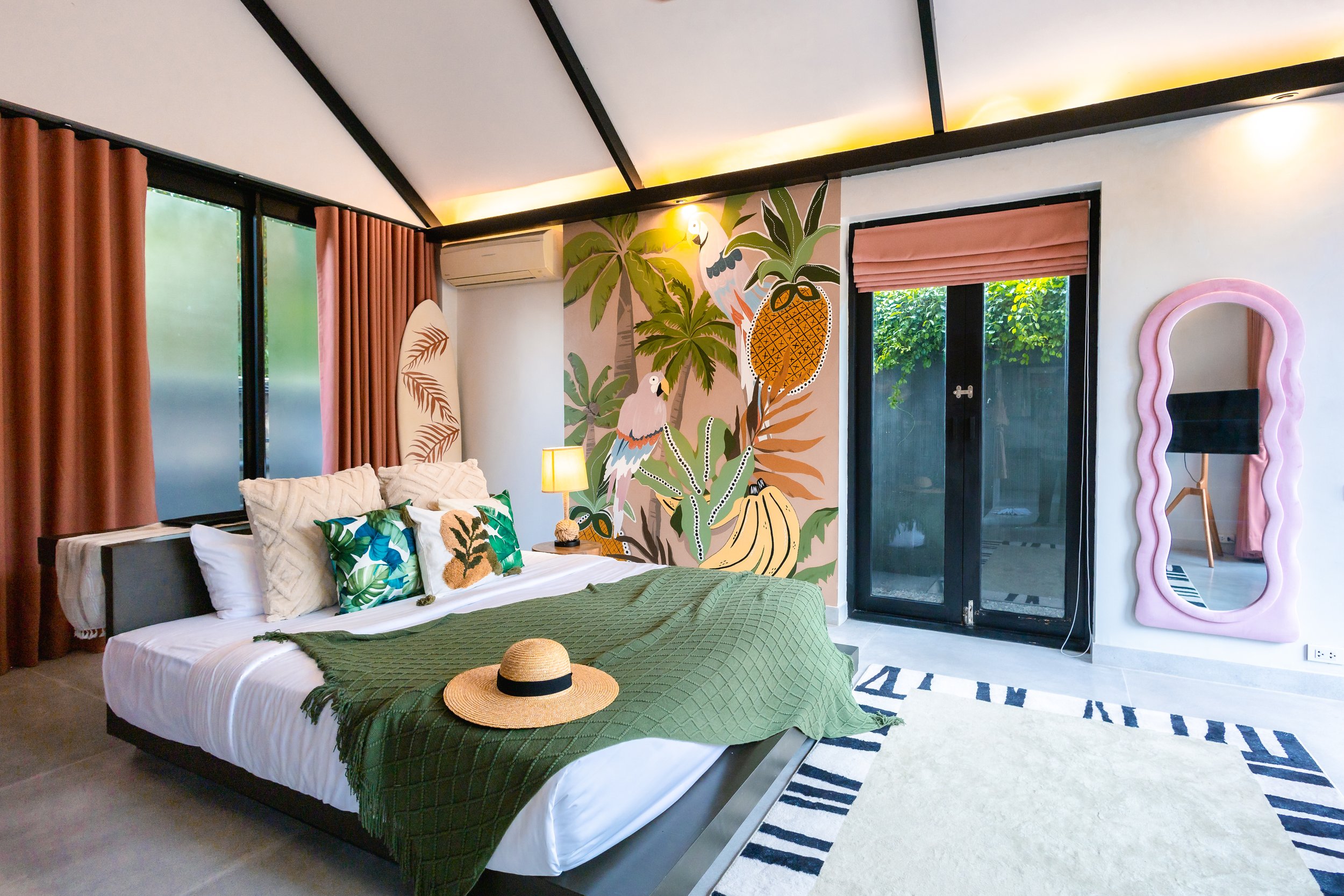 Modern bedroom with tropical-themed wall mural, large bed with green quilt, multiple pillows, pink curtain on window, glass door, pink mirror frame, and a rug.
