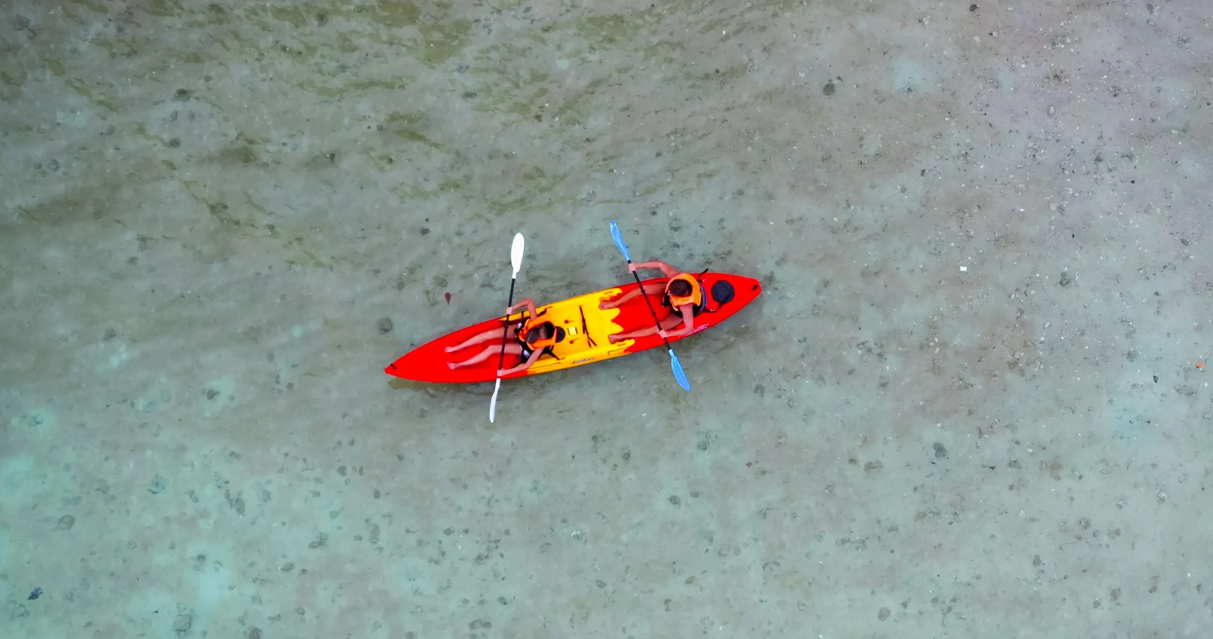 Top-down view of a red and yellow kayak with two people wearing orange life jackets paddling on a sandy surface.