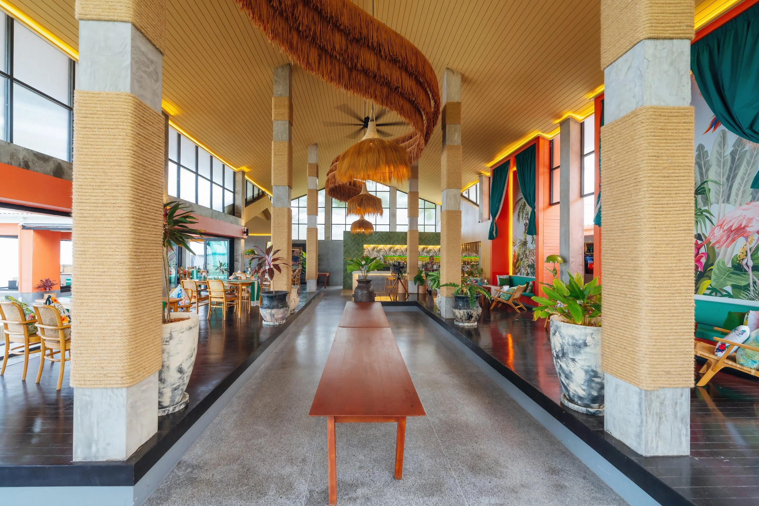 Interior of a bright, tropical-themed restaurant or lounge with large windows, vibrant green plants in large pots, colorful curtains, wooden furniture, and hanging bamboo pendant lights.