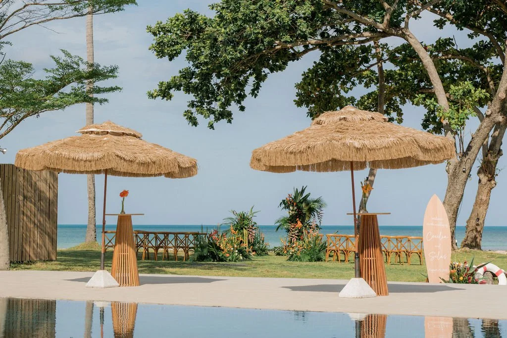 Two thatched umbrellas near a pool with a tropical beach in the background, including palm trees, flower arrangements, and wooden lounge chairs
