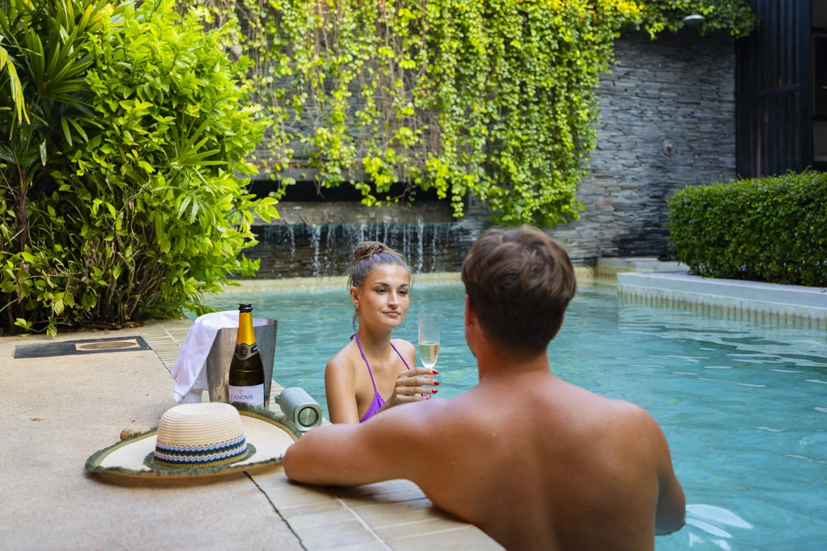 A young woman and man relaxing in a swimming pool, with the woman holding a glass of champagne, surrounded by lush greenery and a stone wall.