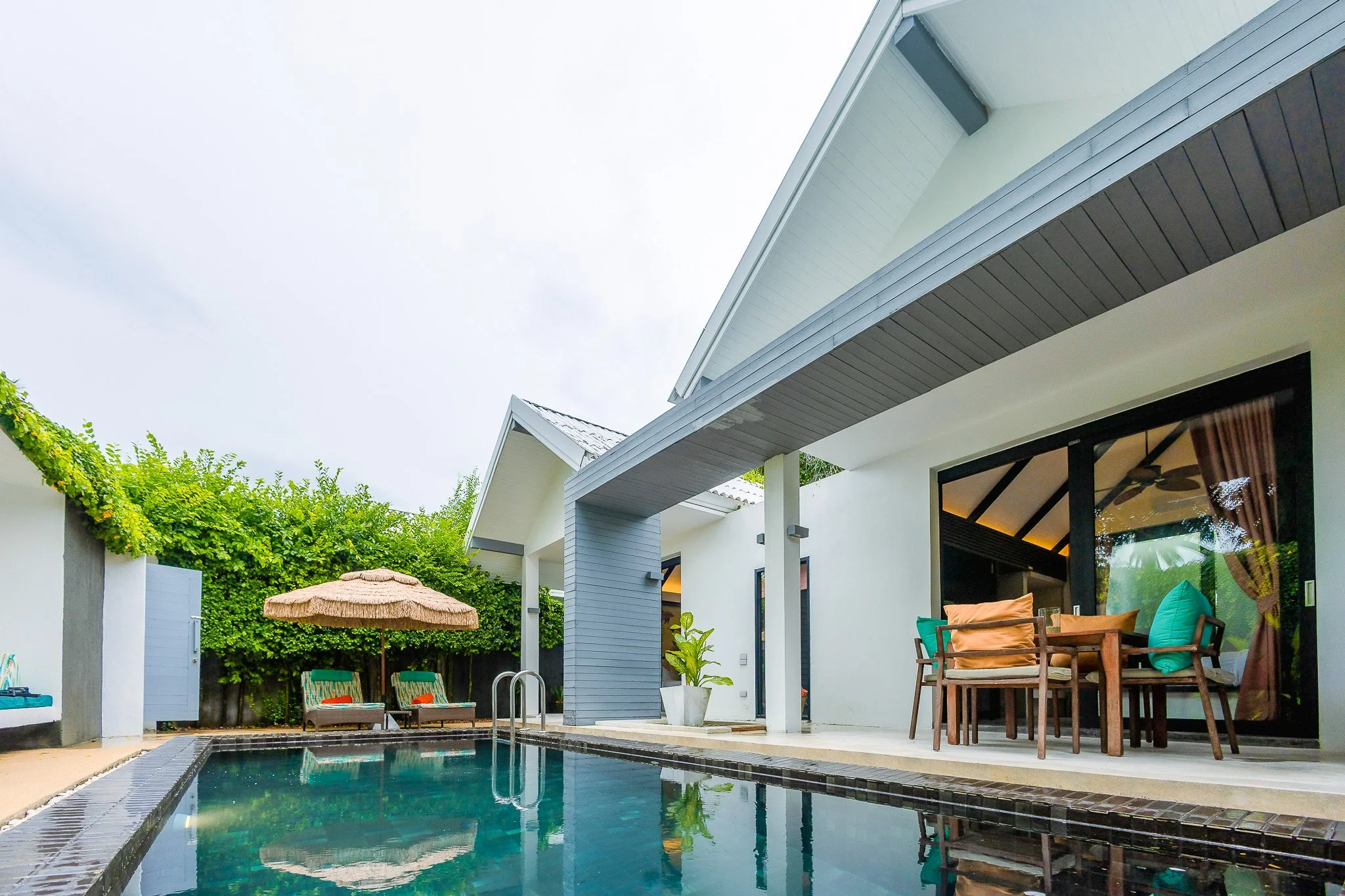 Contemporary house with a private pool, outdoor dining table with chairs, lounge chairs under a straw umbrella, and lush greenery in the background.