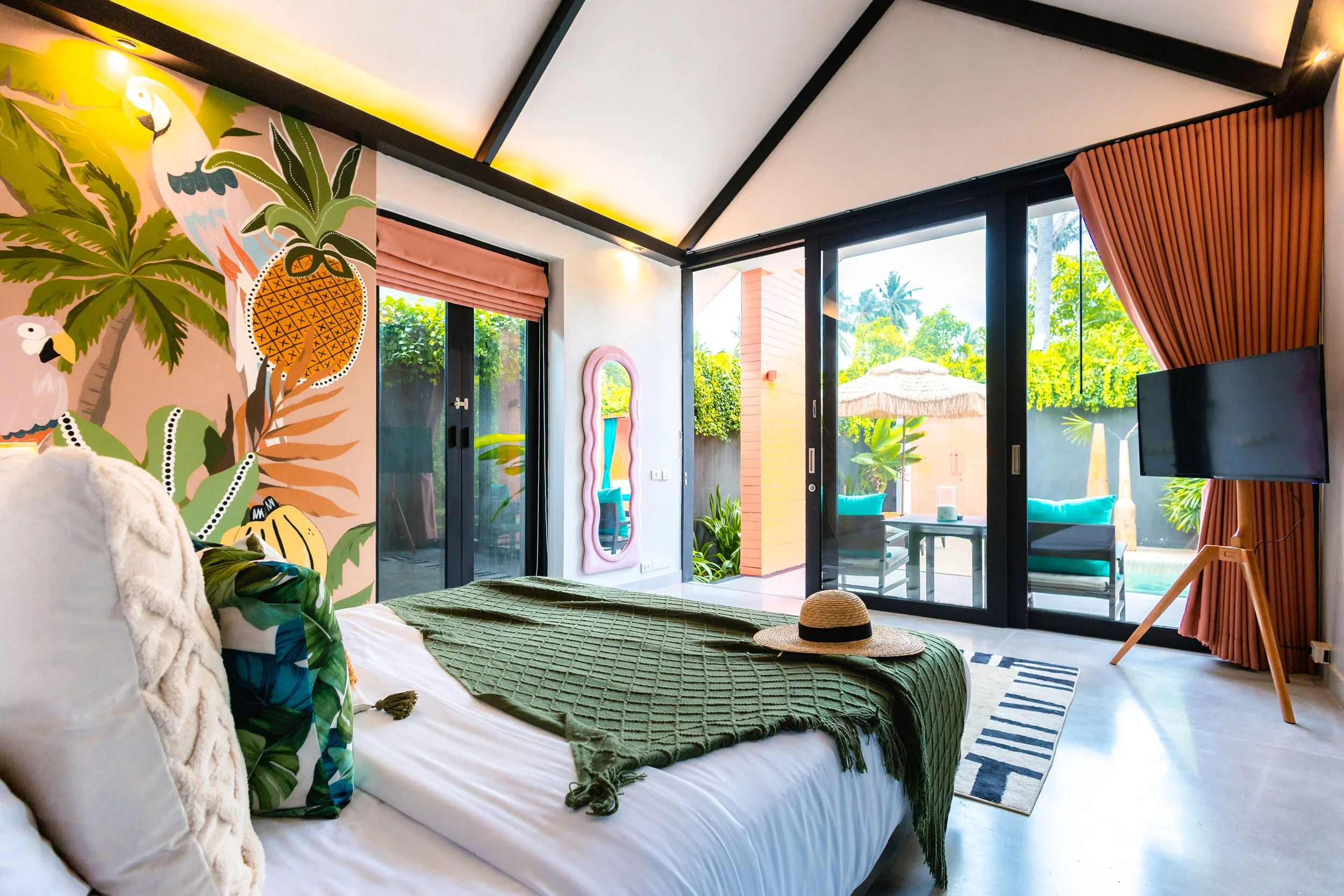 A colorful bedroom with a tropical theme featuring a mural of palm trees, parrots, and pineapples on the wall, a bed with tropical cushions and a green throw blanket, sliding glass doors opening to a patio with outdoor seating and a pool, and vibrant curtains on the windows.