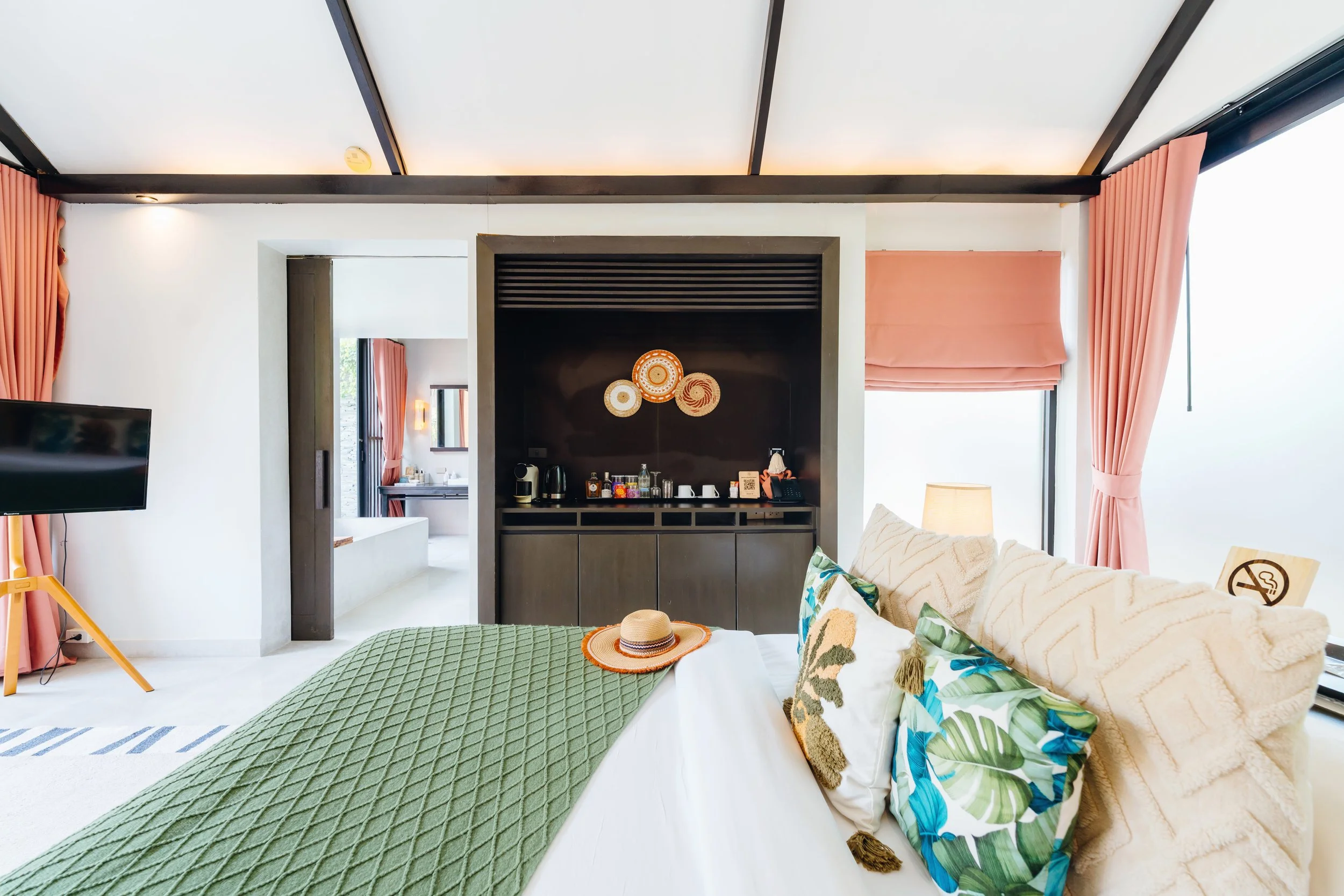 A hotel room with a bed covered in a green quilt, decorative pillows, a sun hat on the bed, a sofa with pillows, pink curtains, and a black built-in cabinet with a coffee station and wall art, bright natural light.