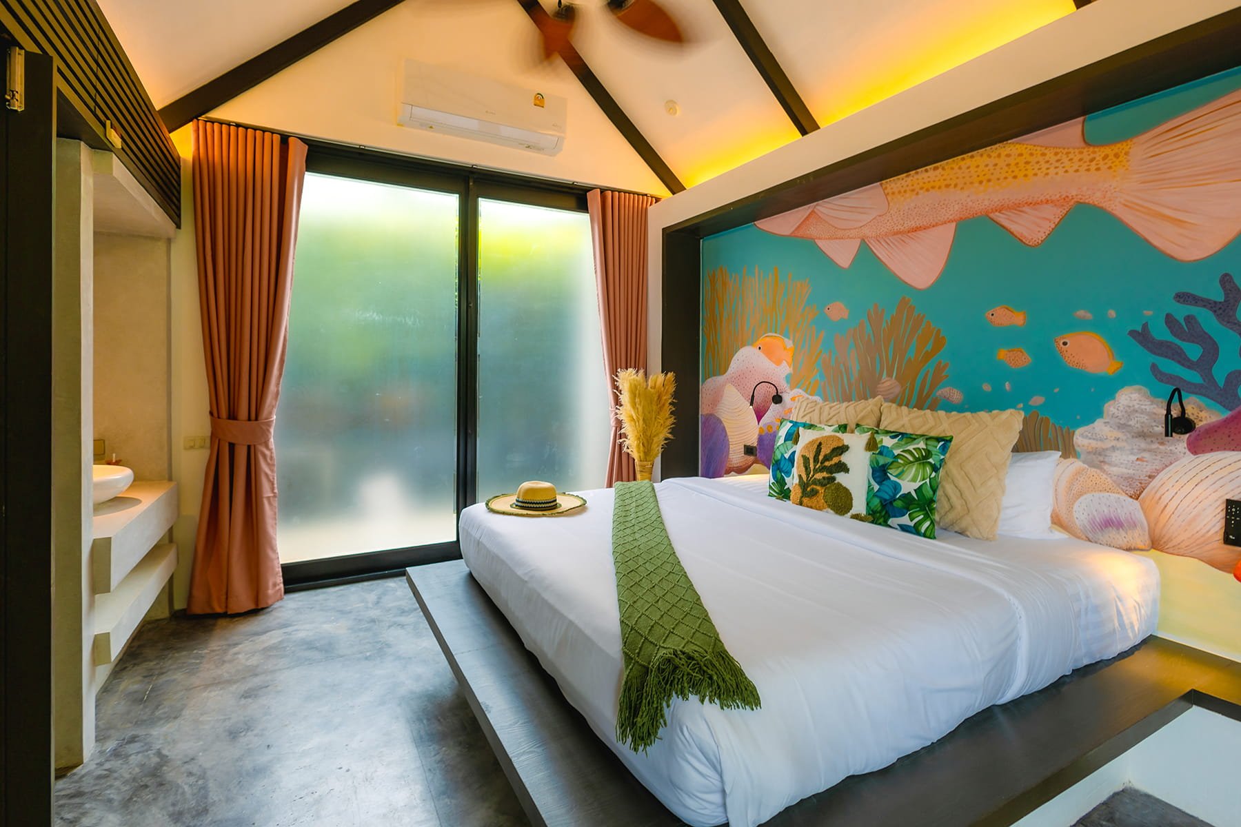 Modern bedroom with aquatic-themed mural wall, large bed with floral pillows, green throw blanket, straw hat on the bed, pink curtains, sliding glass door, and minimalist decor.