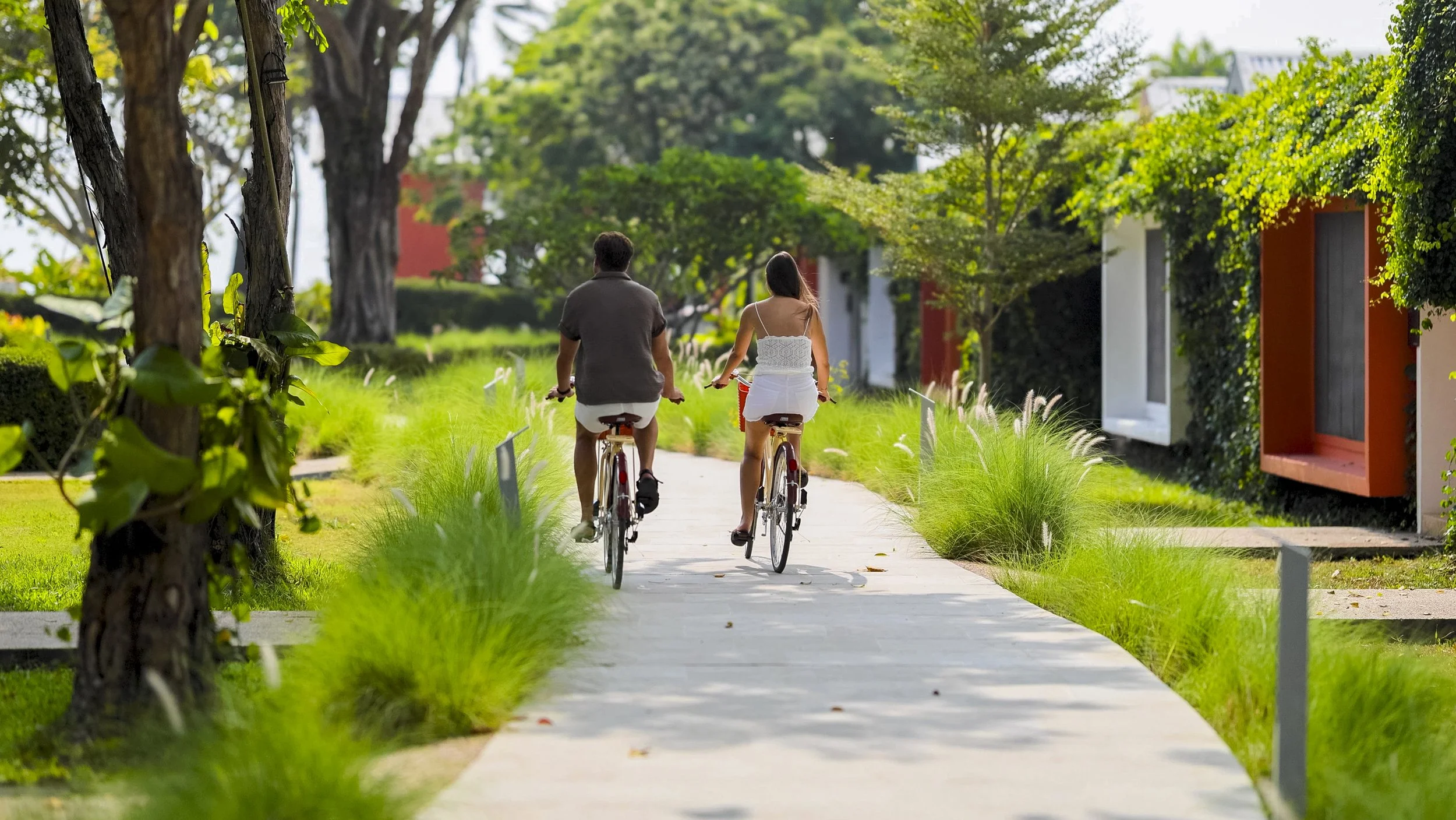 Two people riding bicycles on a paved path surrounded by lush green trees and grass.