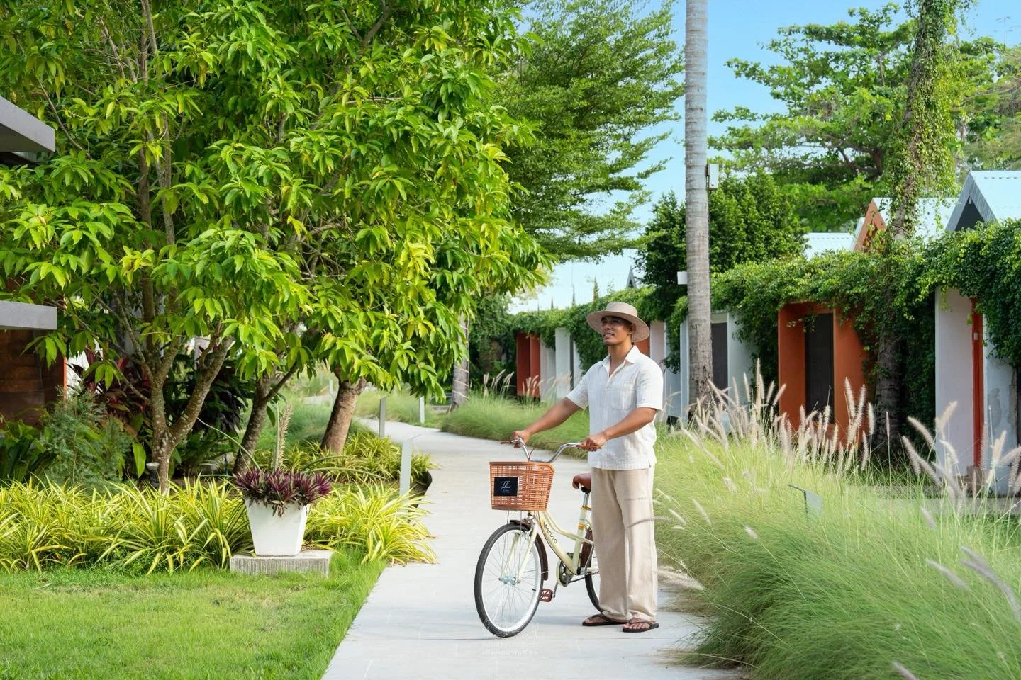 A man in beige pants, a white shirt, sandals, and a wide-brimmed hat stands holding a bicycle with a basket in a lush green outdoor setting with trees, plants, and a paved pathway.