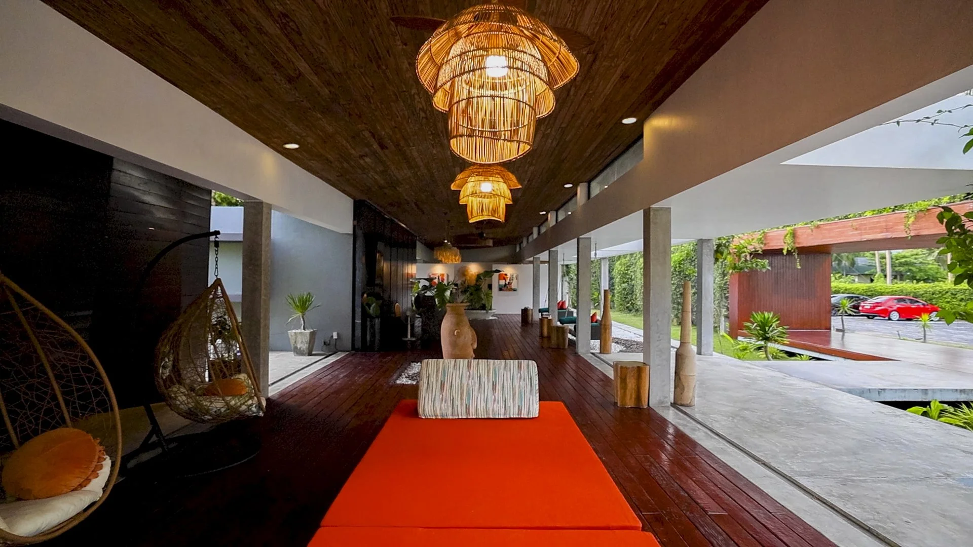 Modern interior with wooden ceiling and orange pendant lights, sitting area with chairs, decorative vases, and lush greenery outside.