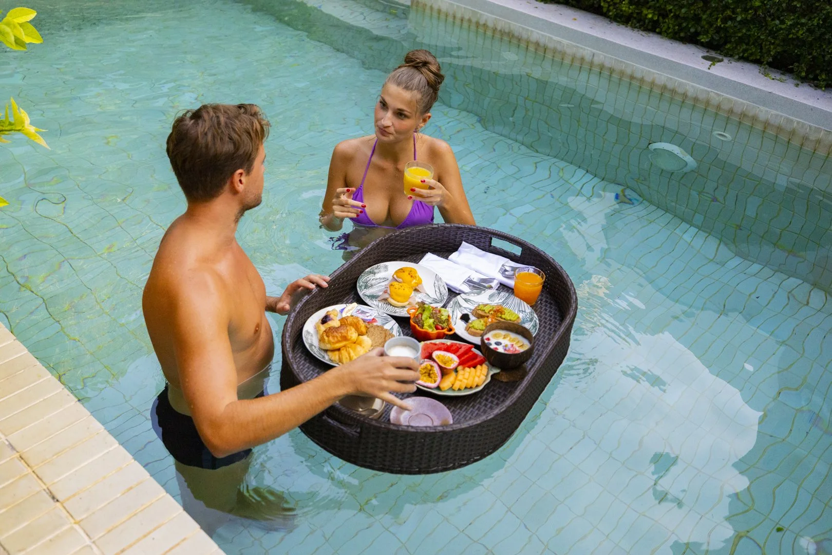 A man and woman in a swimming pool having breakfast together. The woman is wearing a purple bikini and holding a glass of orange juice, while the man is holding a mug. There is a tray with various breakfast foods, including fruits, pastries, and cere