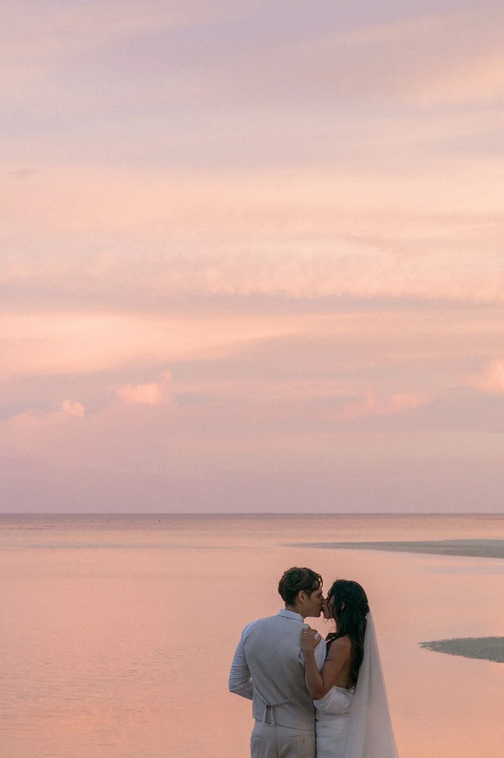 A couple kissing on the beach at sunset, with soft pastel-colored sky and water.