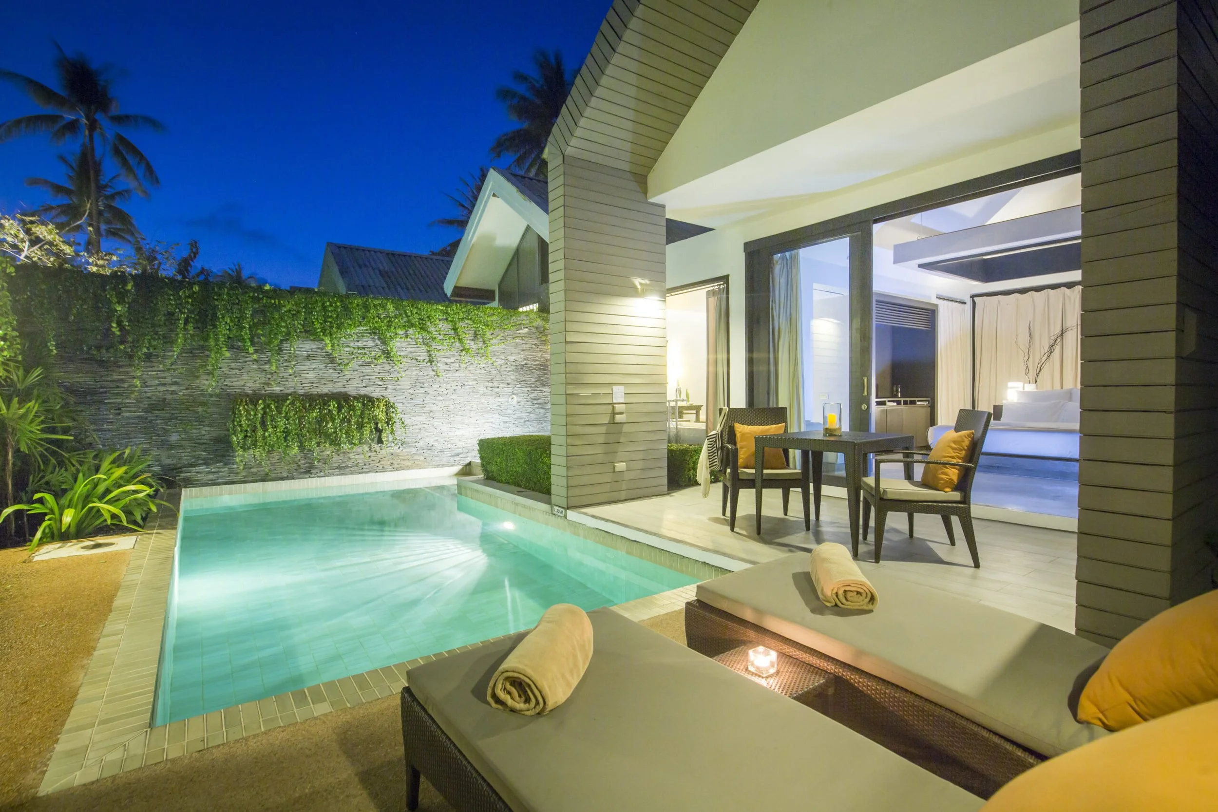 Luxury villa with private pool at night, outdoor lounging area with rolled towels, candles, and an indoor bedroom visible through sliding glass door.