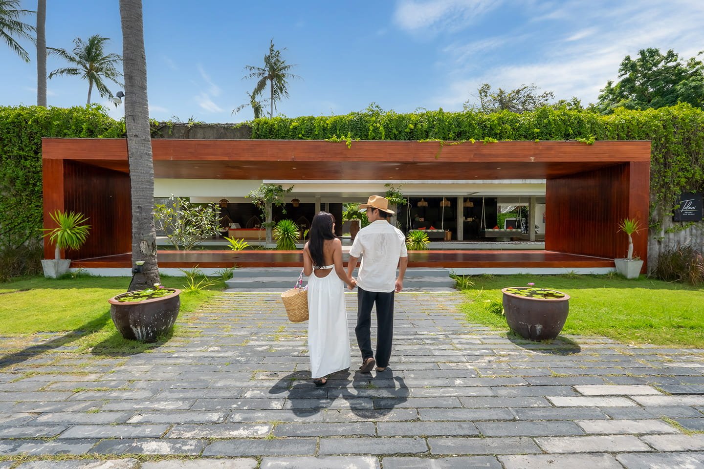 A couple walking hand in hand towards a modern outdoor restaurant or cafe with lush greenery and potted plants, under a blue sky.
