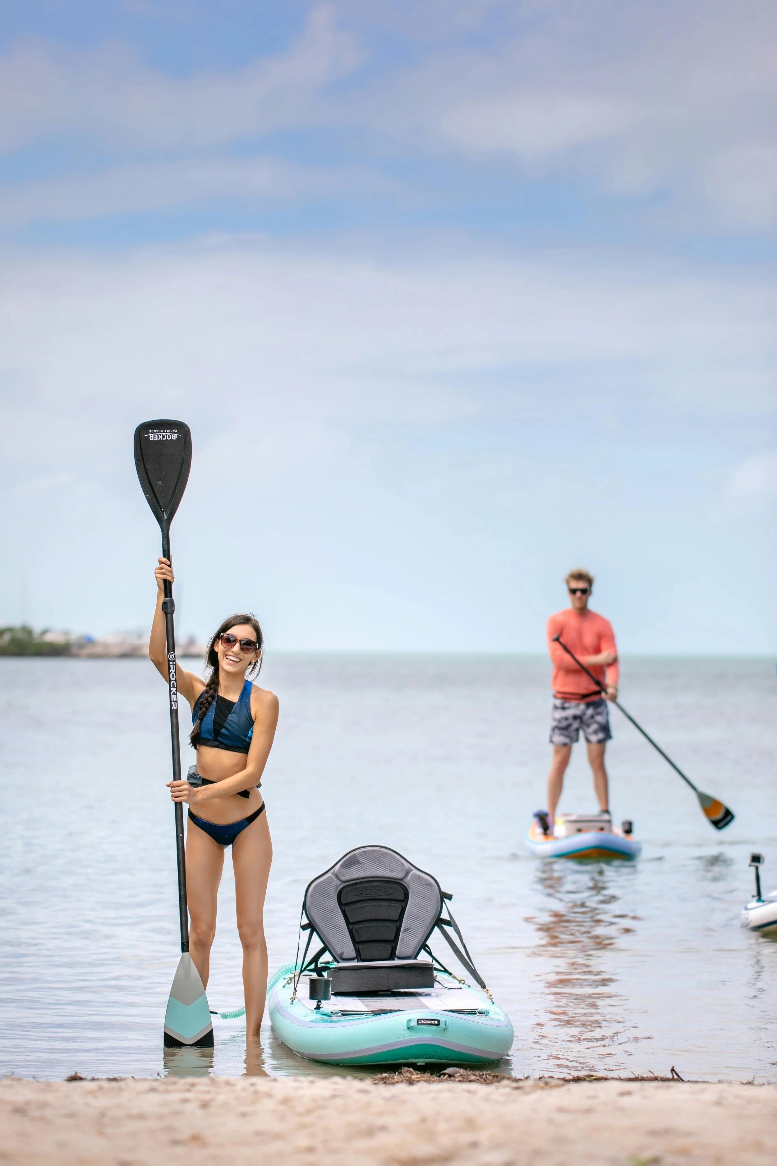 A woman in a bikini holding a paddle next to a paddleboard on the beach, with a man paddleboarding in the background.