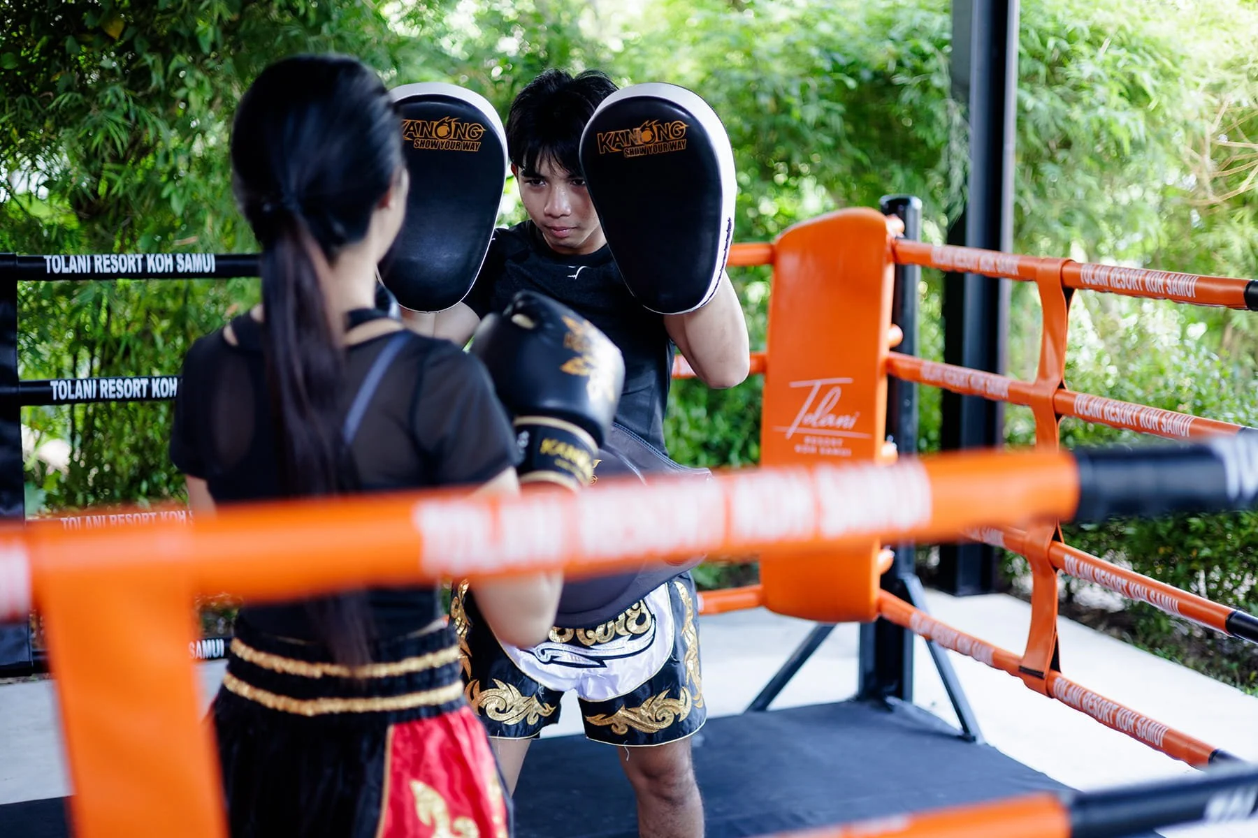 Two Muay Thai fighters practicing in a ring with green foliage background, one man wearing black shorts with gold designs and the other woman in black and red shorts, both wearing black gloves and protective headgear.