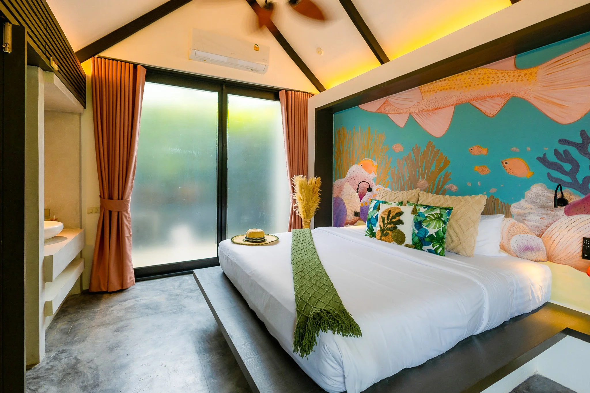A modern bedroom with a colorful ocean-themed mural, a white bed with multicolored pillows, a green blanket, and decorative grasses on a tray, with frosted sliding glass doors and pink curtains.