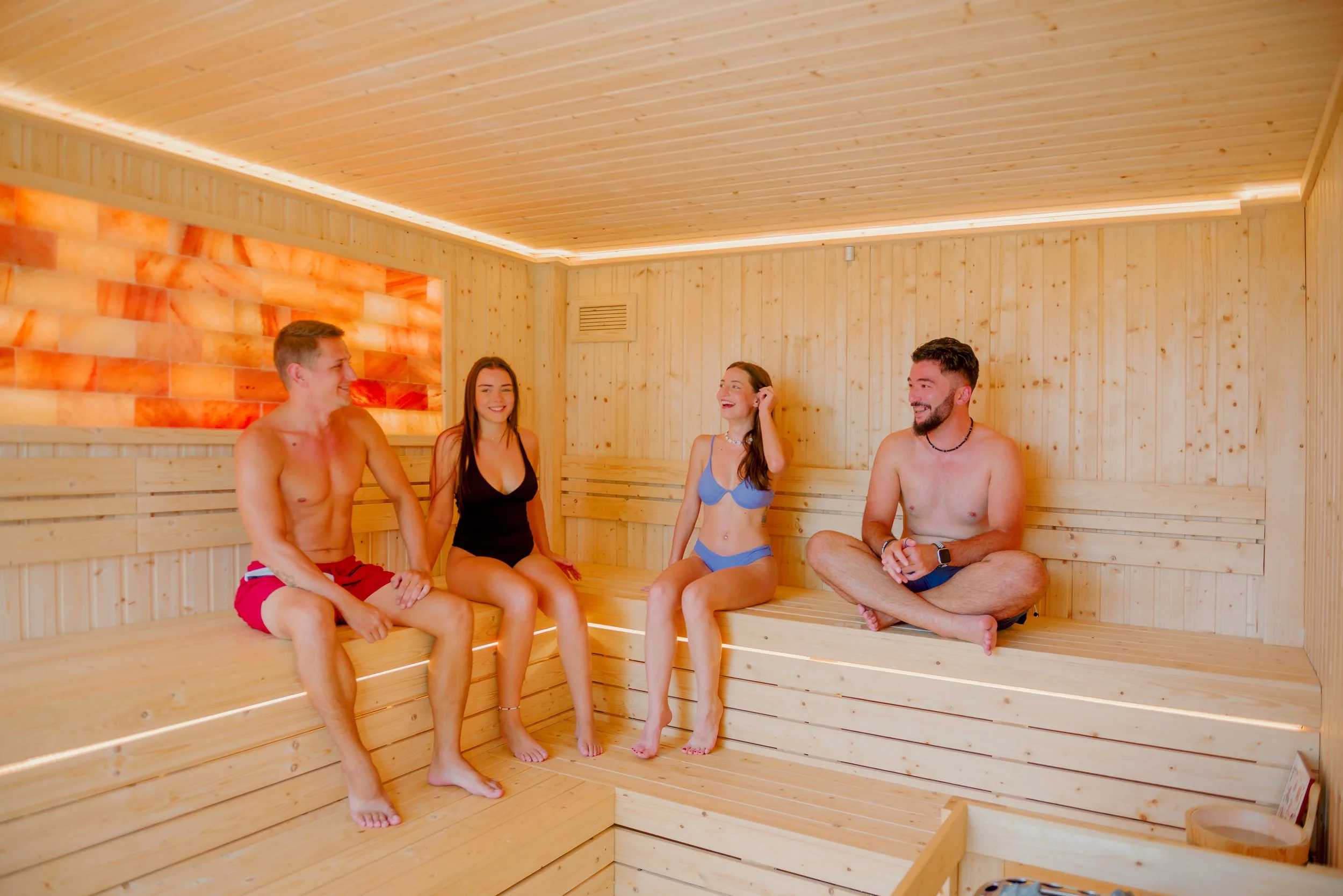 Four young adults sitting inside a wooden sauna, wearing swimsuits, smiling and chatting.