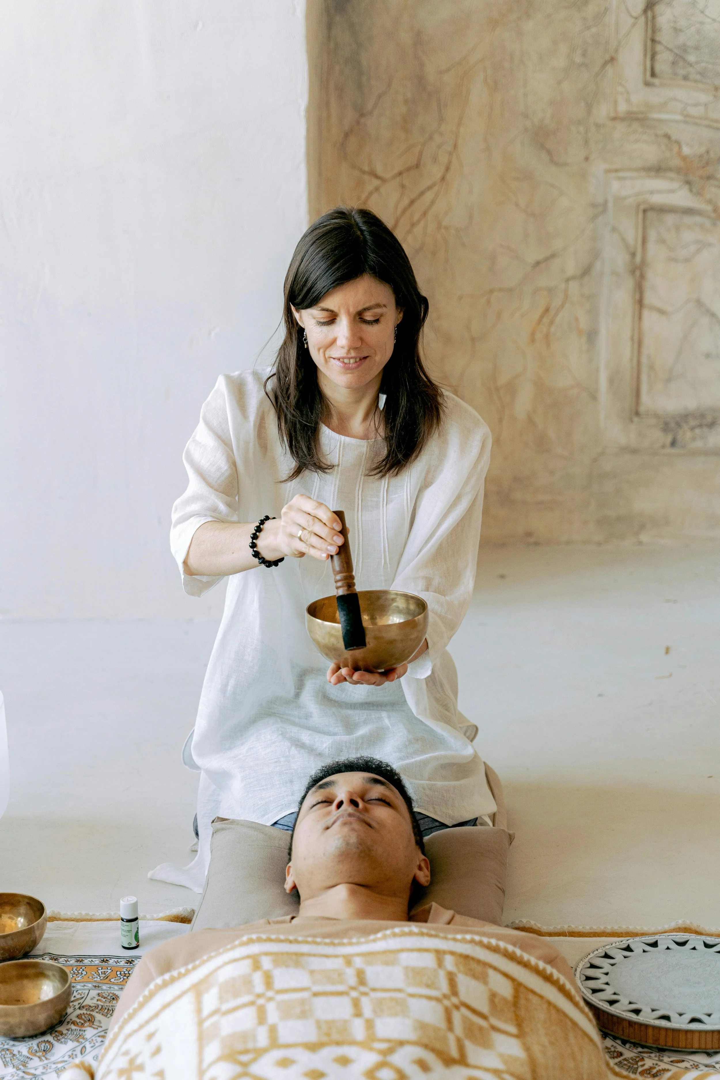 A woman performing sound healing with singing bowls on a man lying down with eyes closed