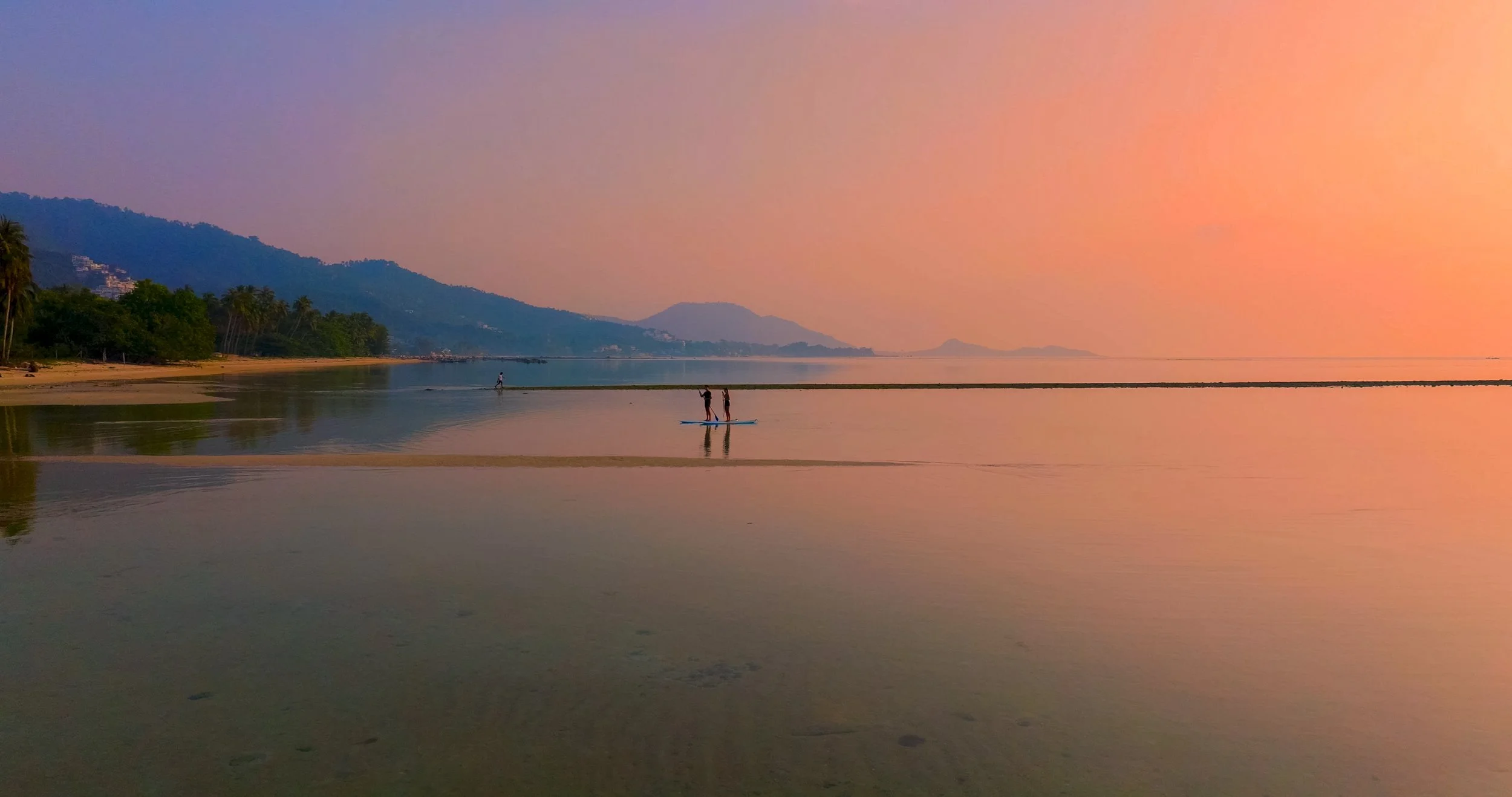 A sunset on a beach with people paddleboarding and walking along the shoreline, with mountains in the background and the sky painted in pink and orange hues.