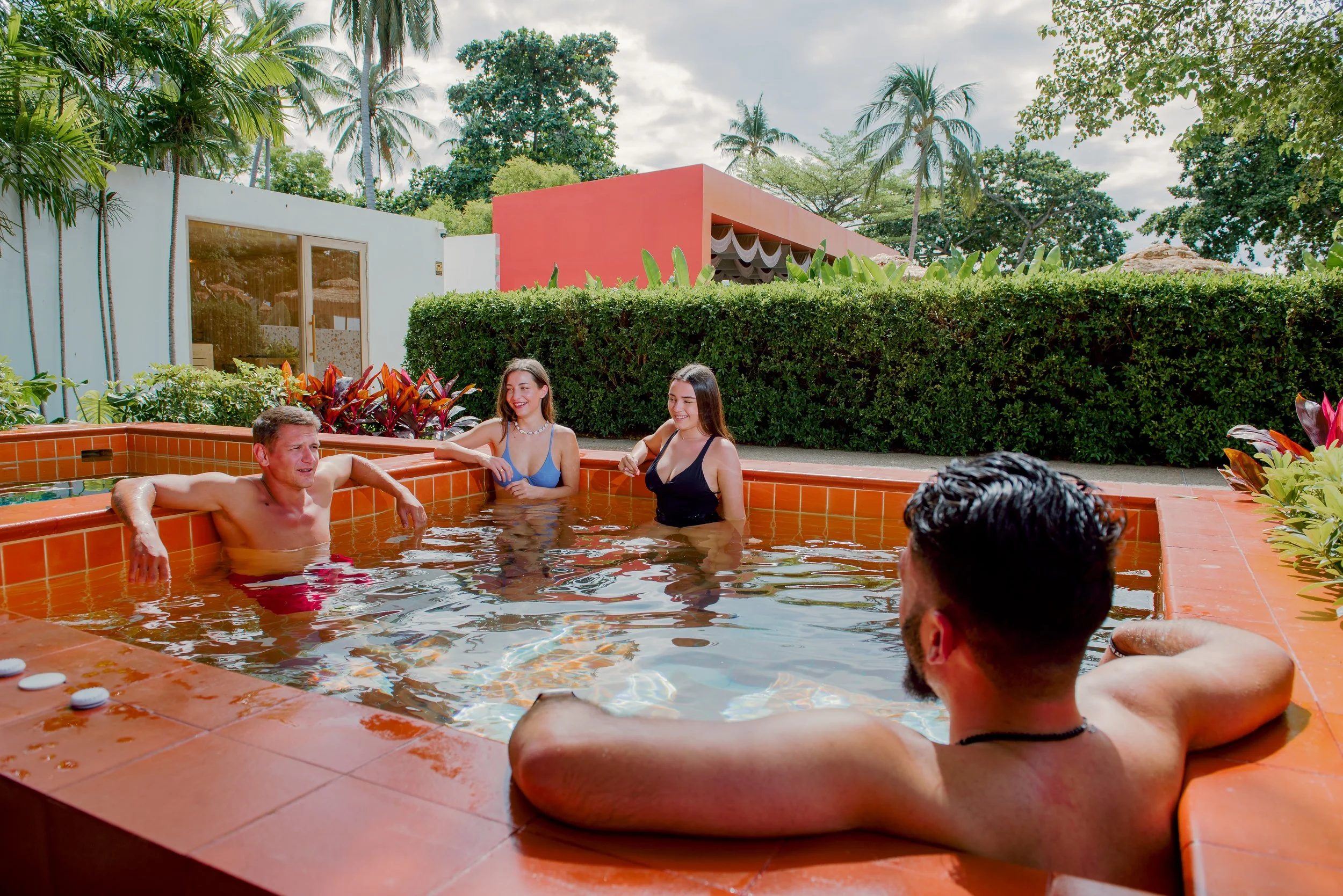 Five friends enjoying a relaxing moment in a hot tub outdoors, surrounded by lush greenery and modern buildings in the background.