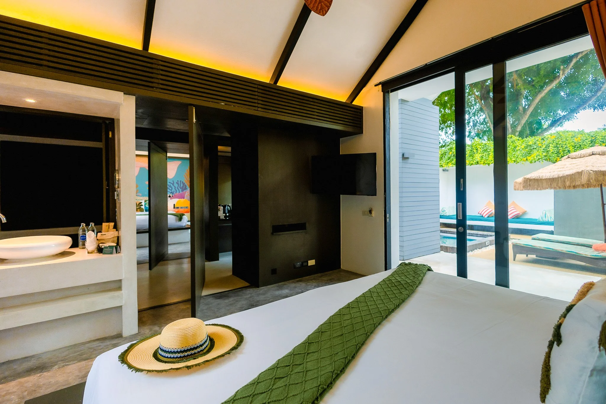 Modern hotel room with large sliding glass door leading to outdoor pool and lounge area, bed with white linens and green runner, straw hat on bed, sea-themed artwork, small sink area, and outdoor umbrellas.