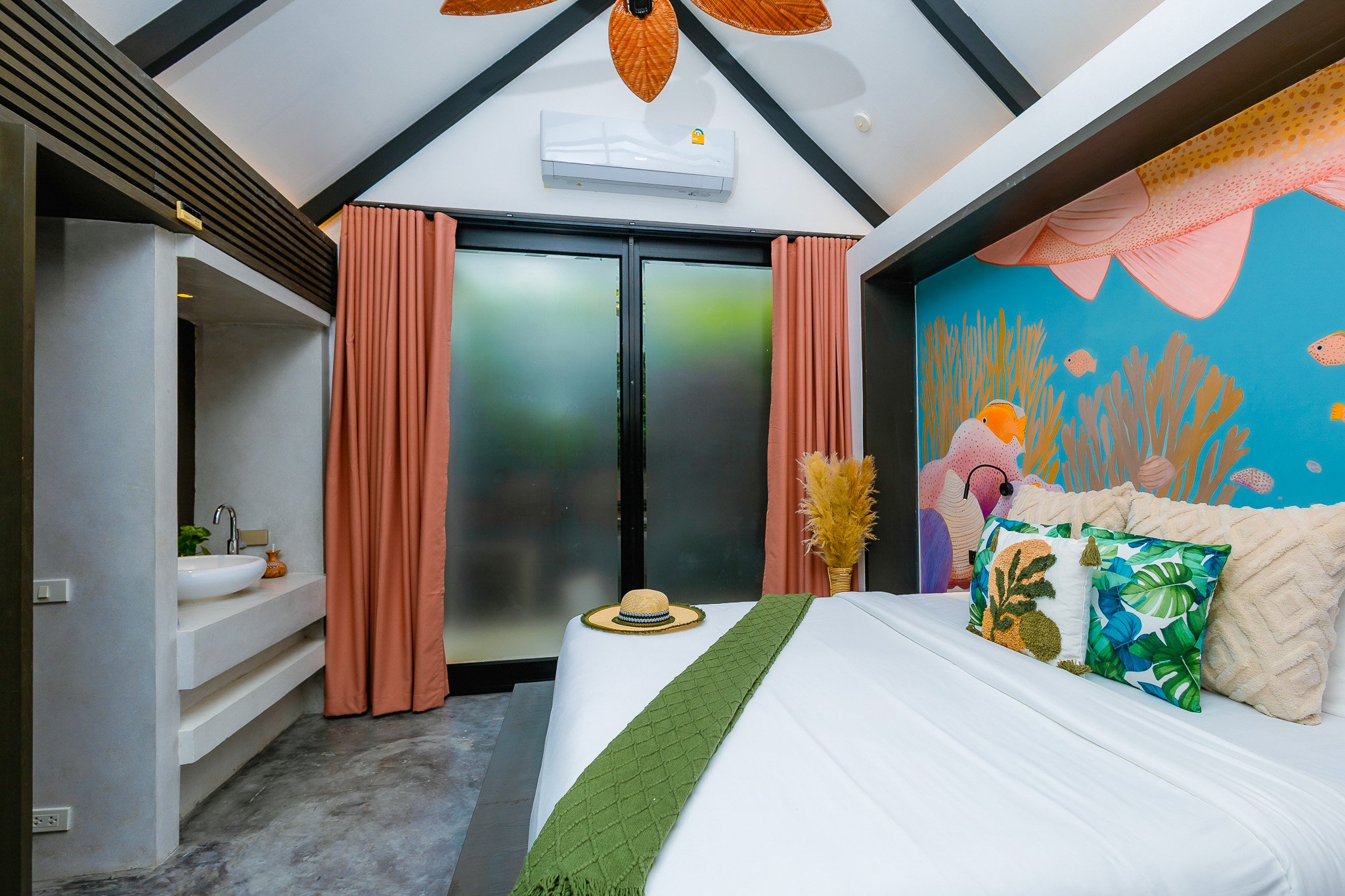A bedroom with a tropical-themed mural on the wall, a white bed with colorful pillows, a green throw blanket, a straw hat on the bed, pink curtains, a small sink area, and a door with frosted glass.