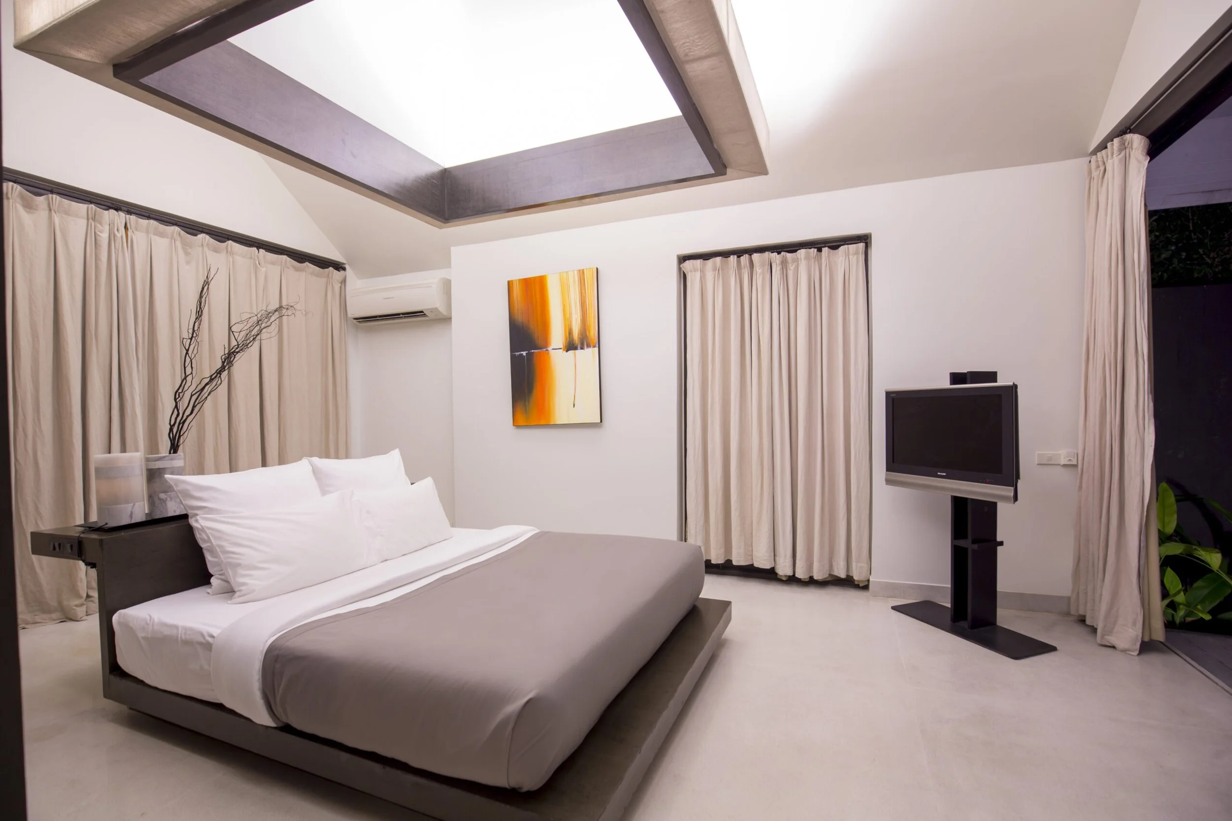 Modern bedroom with a large bed, white bedding, beige curtains, wall art, and a flat-screen TV.