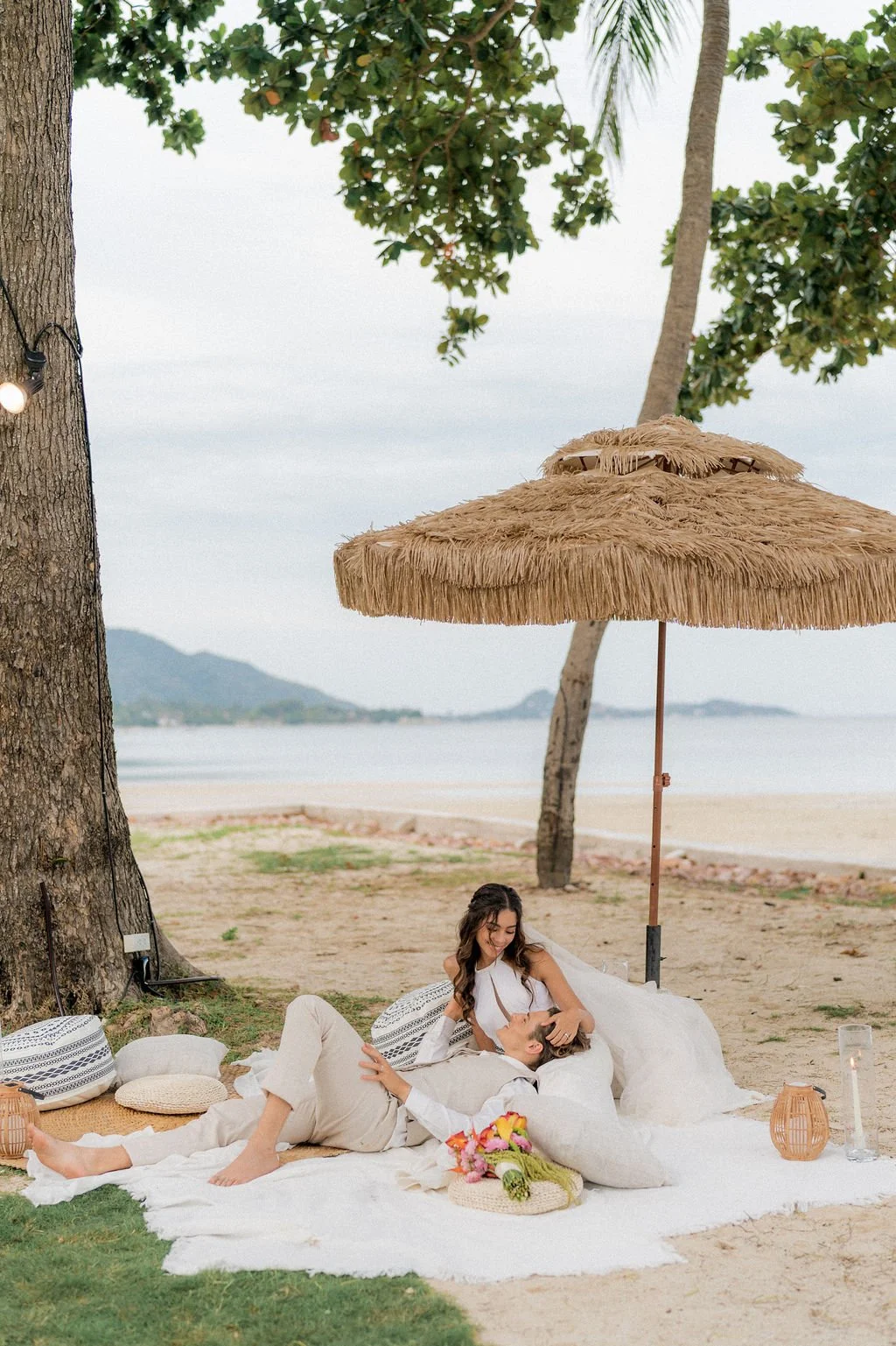 A couple relaxing on a beach under a large straw umbrella with pillows, flowers, and lanterns, with trees and the ocean in the background.