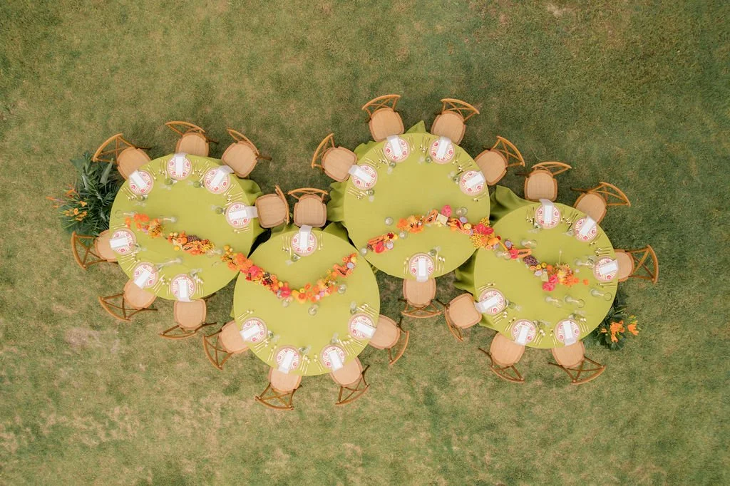 Top-down view of four round green tables with tan chairs, decorated with orange and pink floral arrangements, set on grass for an outdoor event.