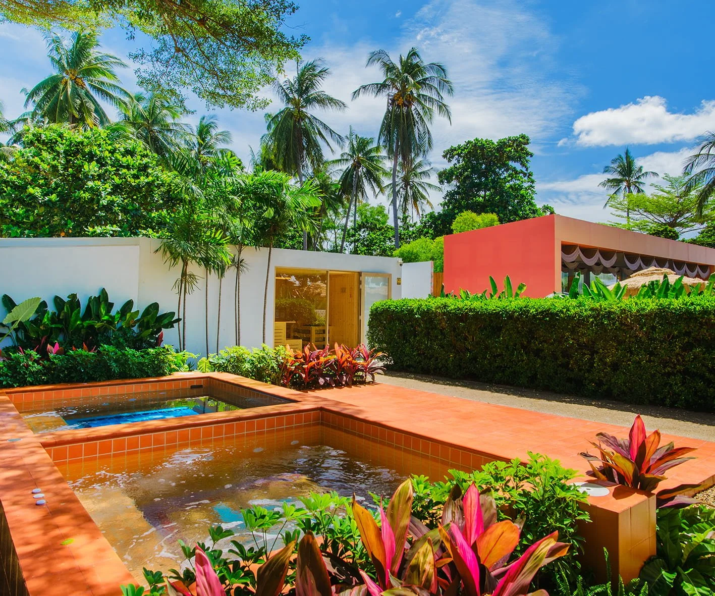 Tropical resort with small hot tubs, lush green plants, tall palm trees, and colorful buildings under a blue sky with clouds.