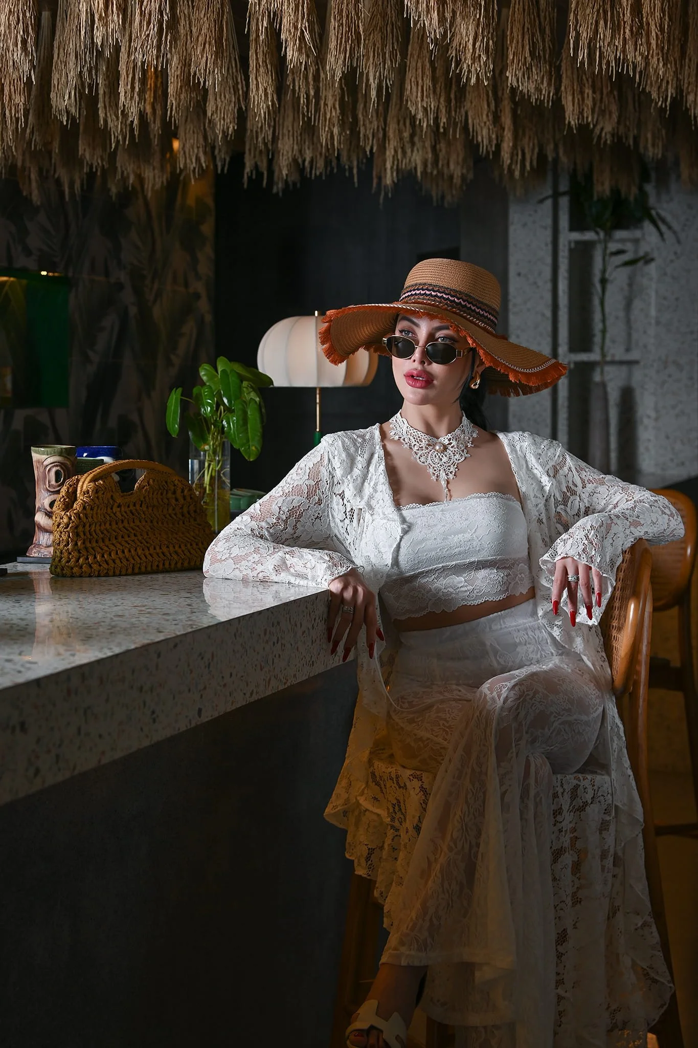 A woman sitting at a bar counter in a trendy, stylish setting, wearing a large sun hat, sunglasses, a white lace off-shoulder outfit, and jewelry.