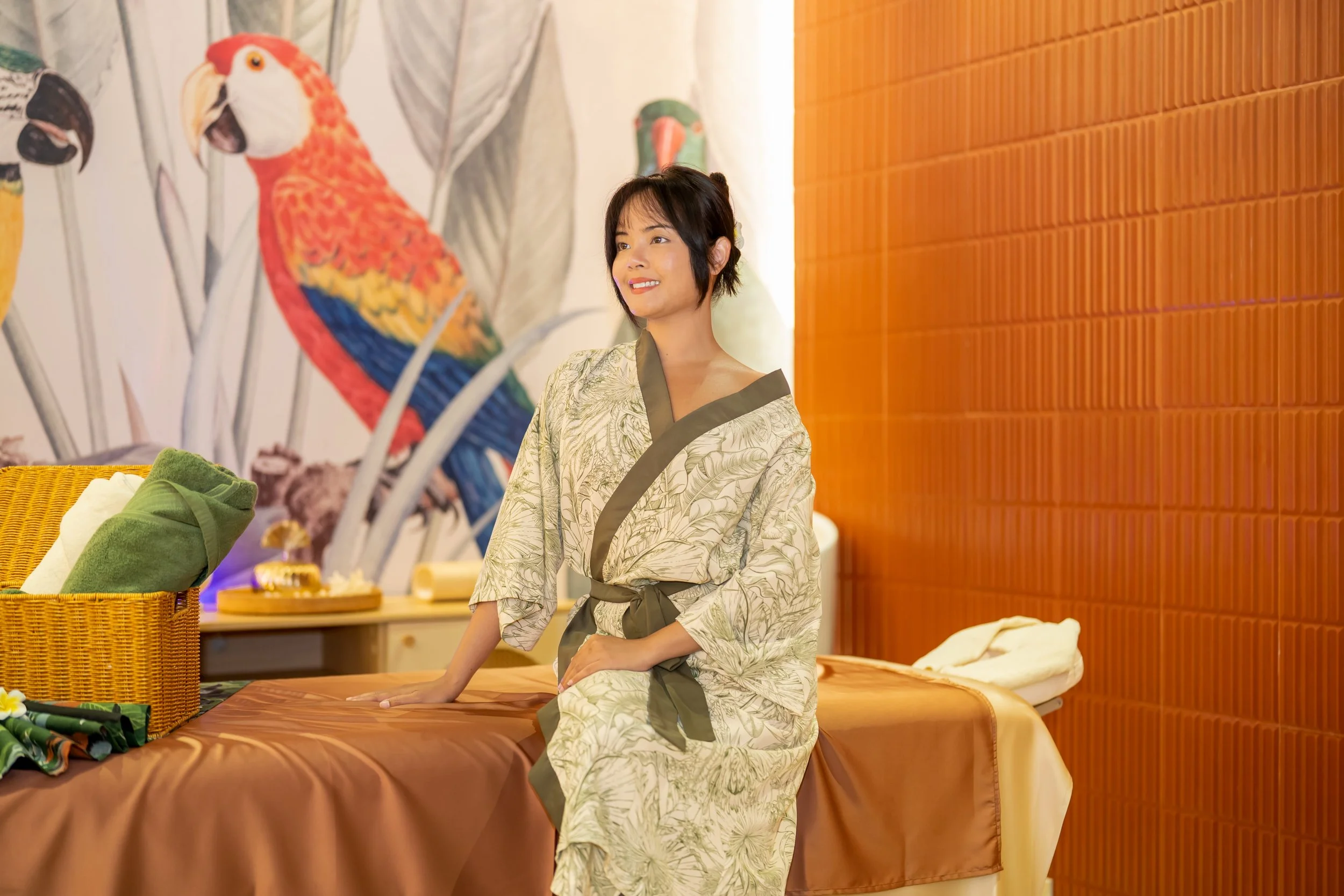 A woman in a patterned bathrobe sitting on a massage table in a spa room with a tropical bird mural on the wall.