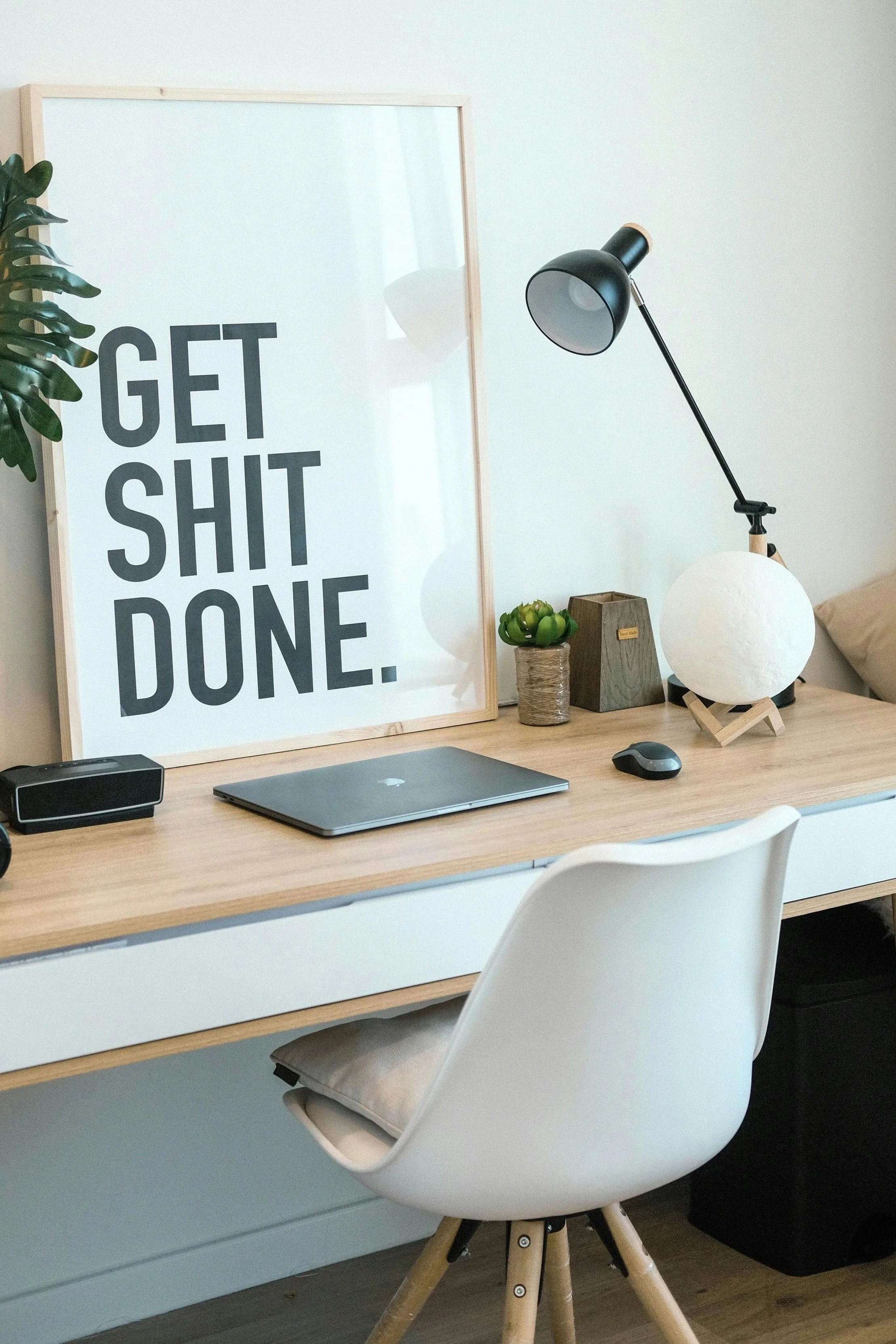 Home office desk with a poster that reads 'Get Shit Done,' a laptop, a lamp, a small plant, and decorative items.