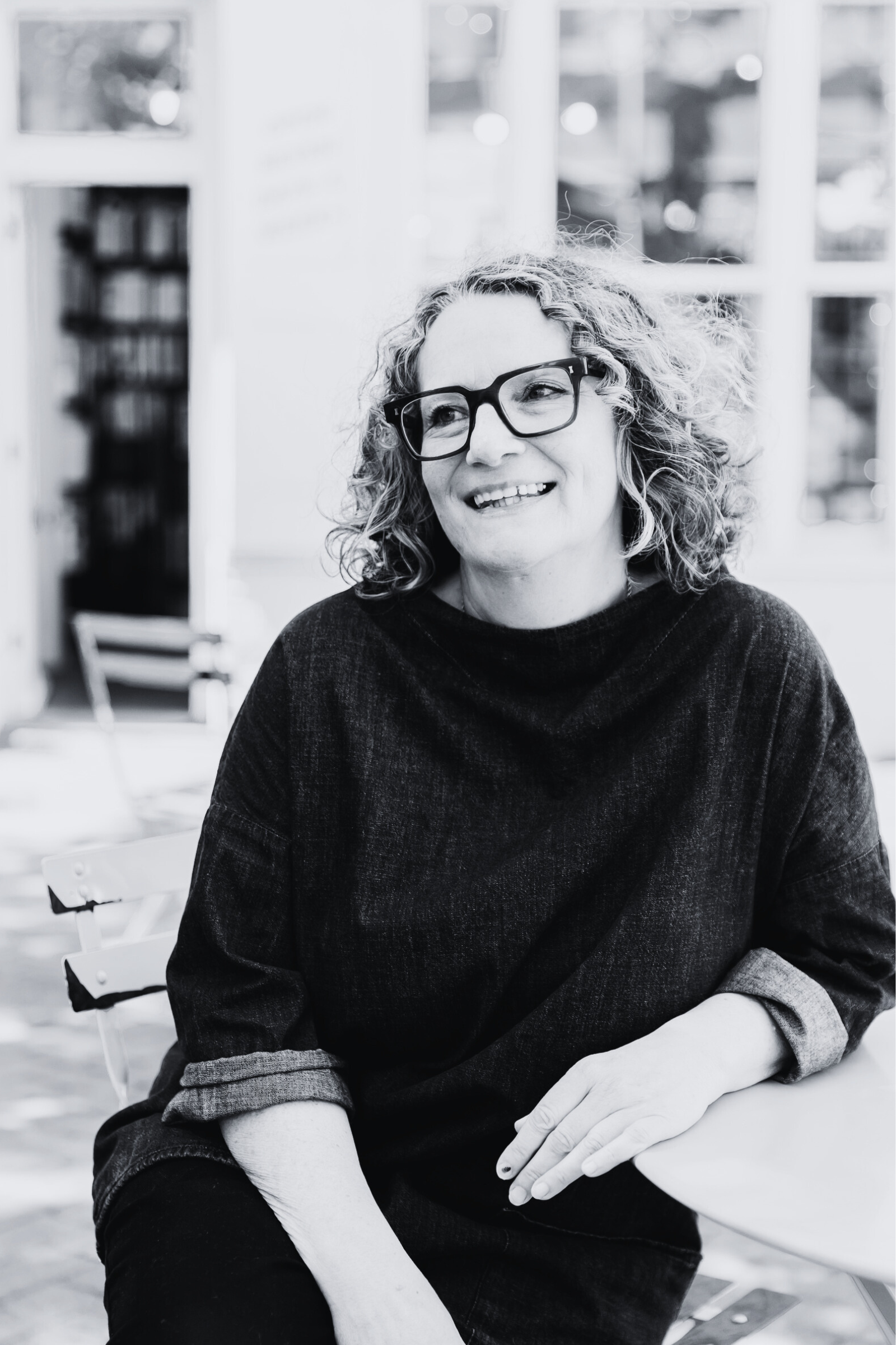 A woman with curly hair wearing glasses, sitting at a table in a bright room, smiling and looking to the side.