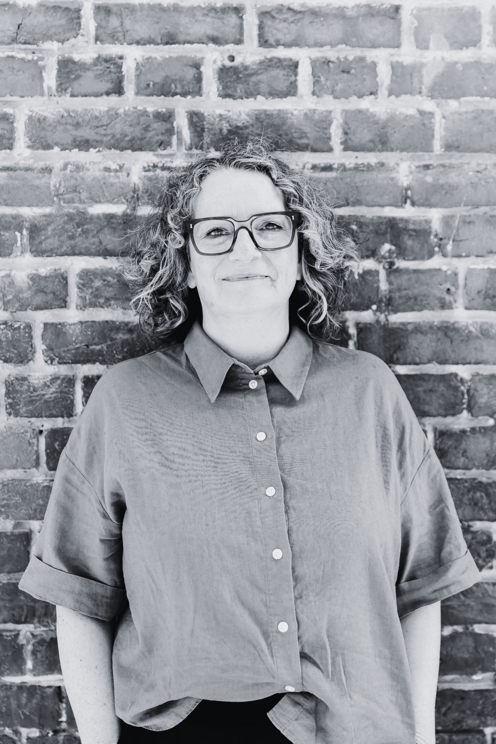 Black-and-white portrait of a woman with curly hair, wearing glasses and a button-up shirt, standing against a brick wall.