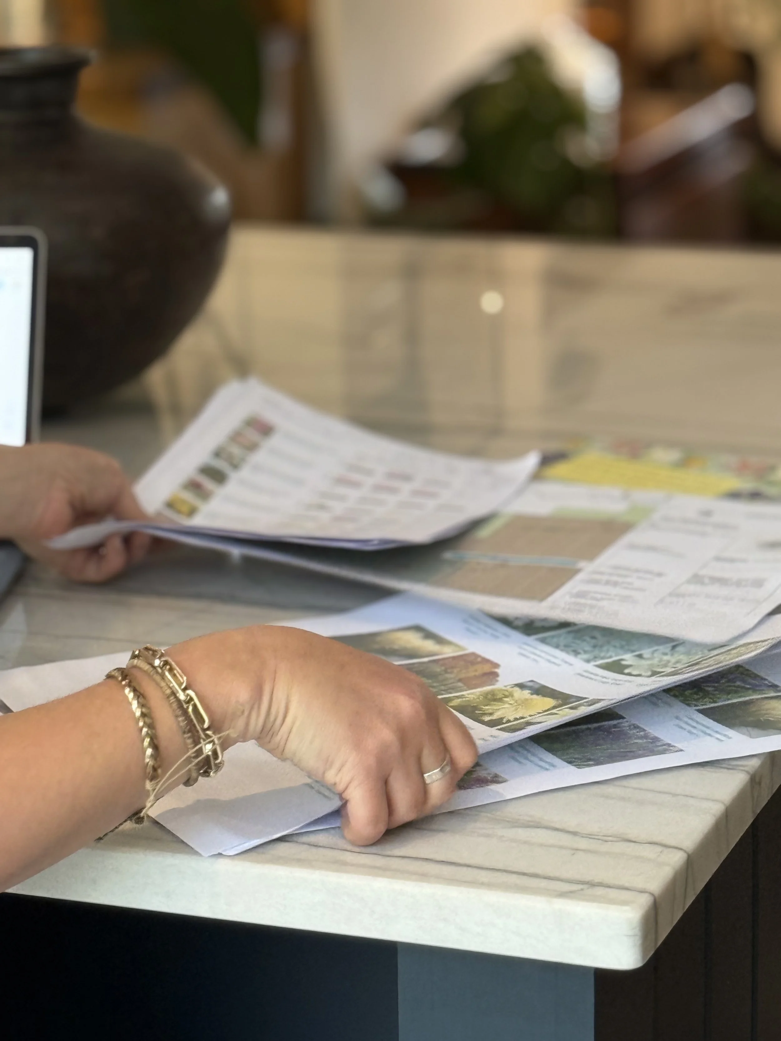 A person with bracelets on their wrist is flipping through printed papers or brochures on a light-colored table, with a large dark vase in the background.