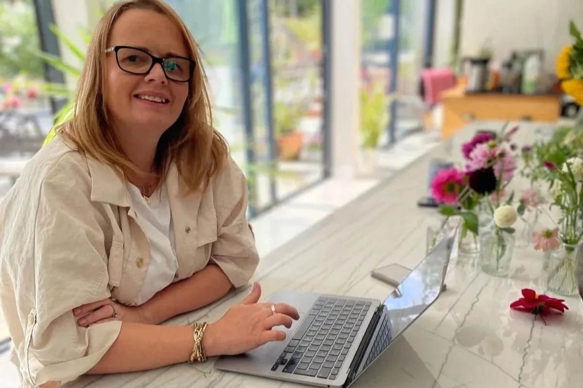 Madeline with glasses and light brown hair sitting at a marble table with a laptop, in a bright room with large windows and a view of outdoor plants and flowers.