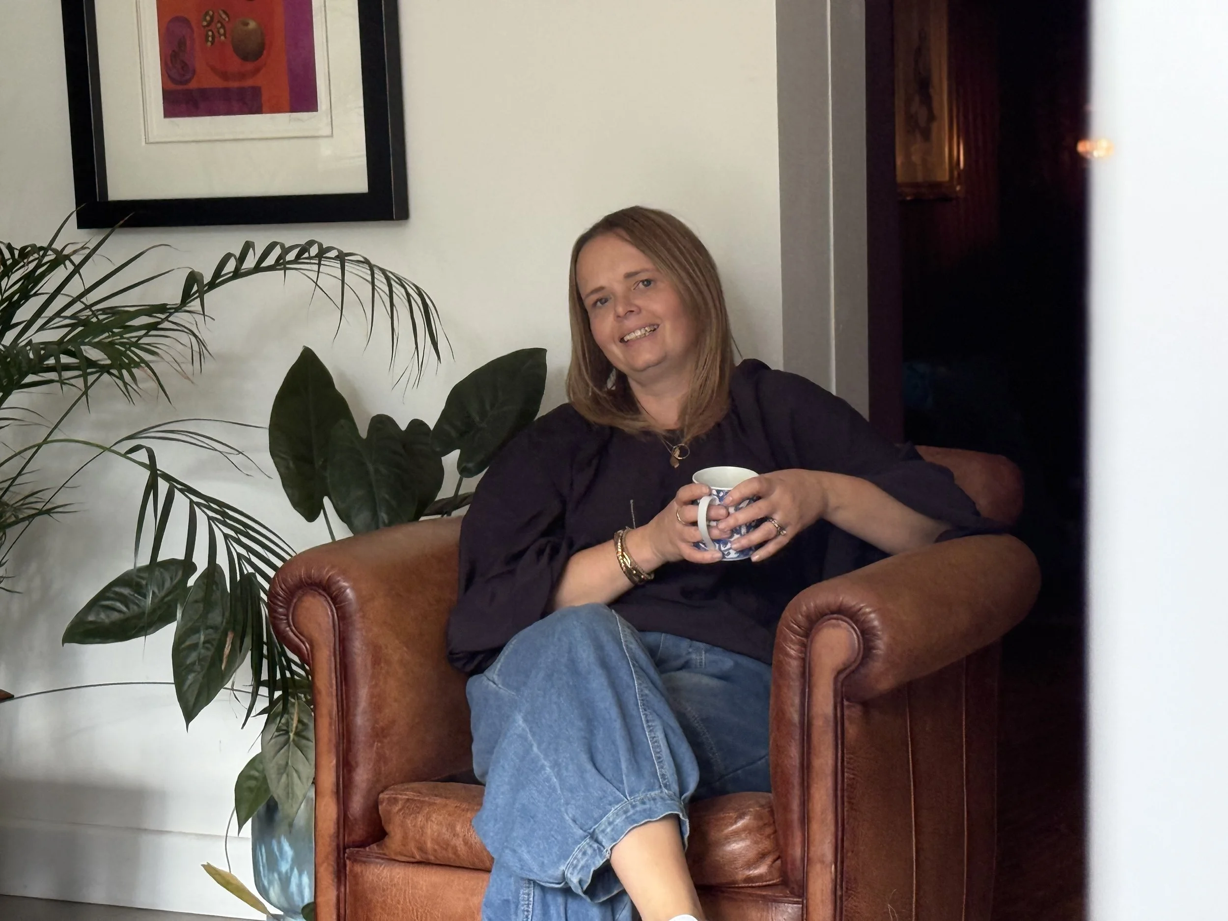 A woman with shoulder-length brown hair smiling and holding a mug while sitting in a brown leather armchair. There are large green plants behind her and a framed colorful artwork on the wall. The setting appears to be a cozy indoor space.